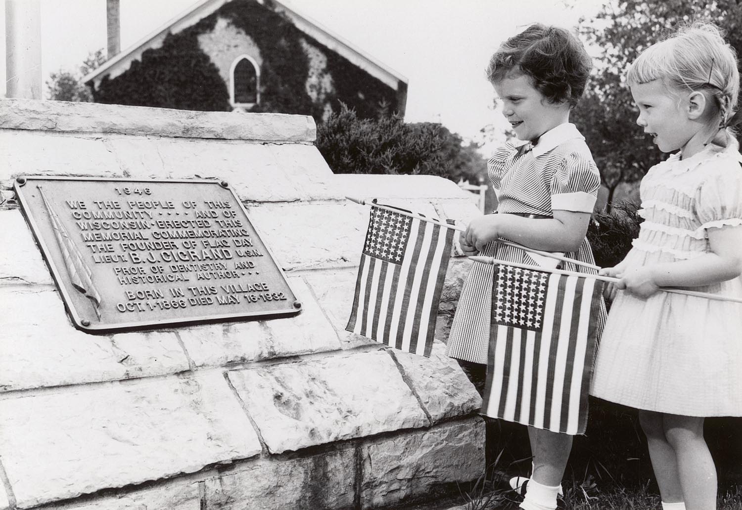 Two young girls hold American flags next to a plaque honoring Bernard J. Cigrand in Waubeka