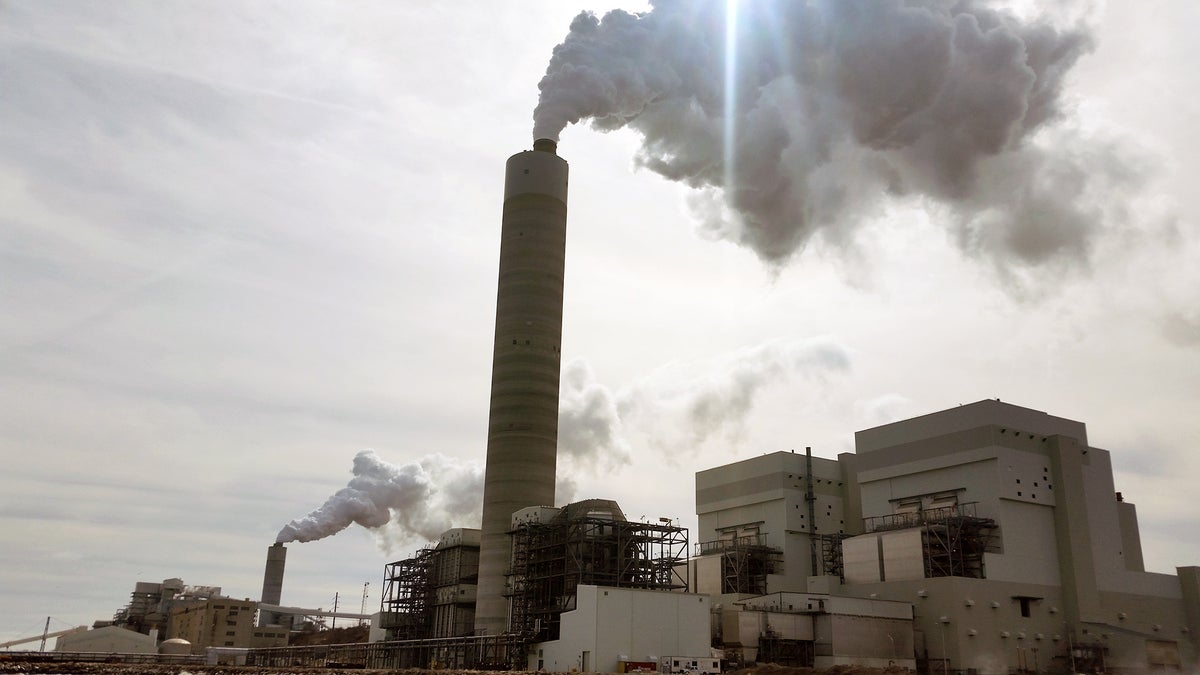 A large industrial power plant emits thick white smoke from tall chimneys into a cloudy sky during the daytime.
