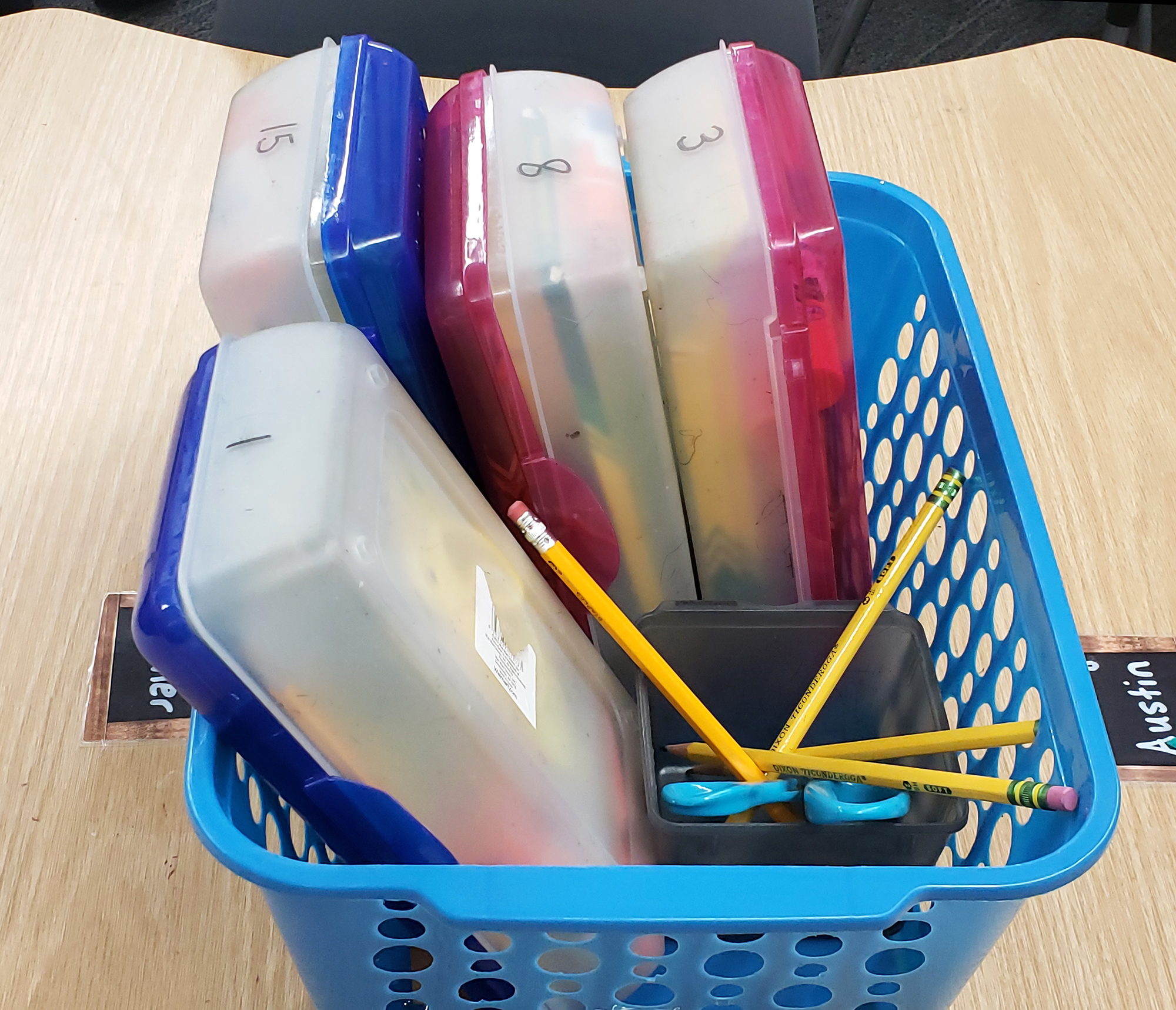 Containers on desks shared by four students each display a number assigned to each student with school supplies such as colored pencils, erasers and glue sticks.