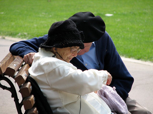An elderly couple sits together on a park bench