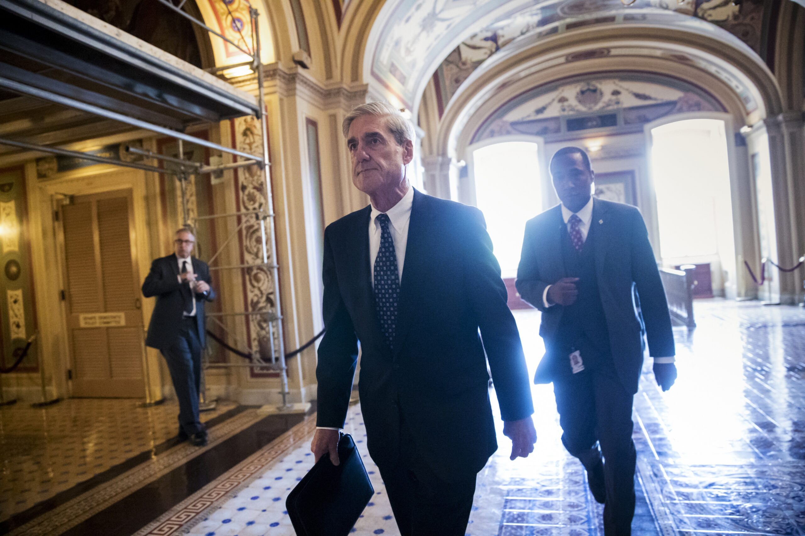 Robert Mueller walking through the Capitol with two men and the sun behind him