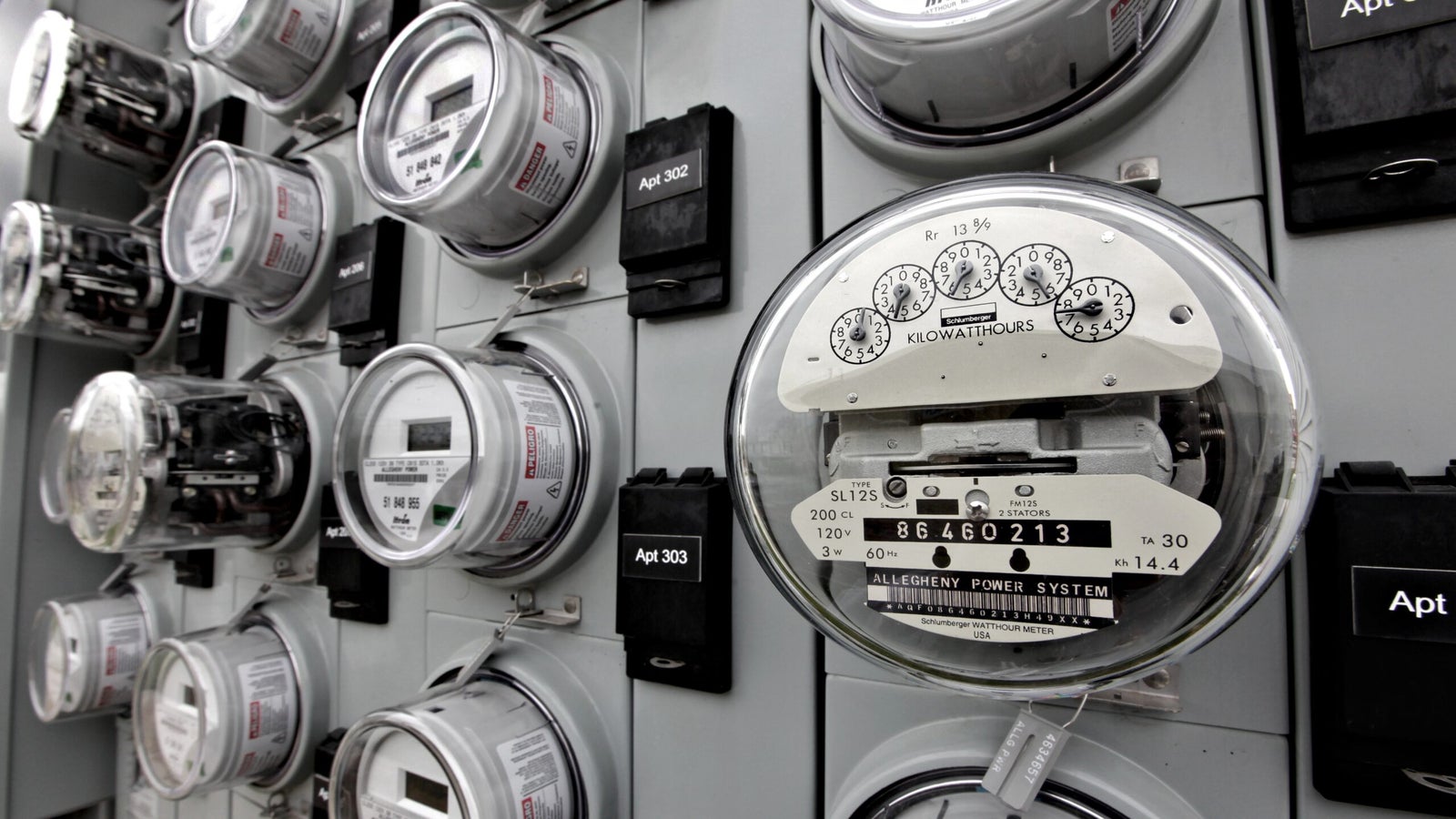 A row of analog electricity meters mounted on a wall, each labeled by apartment number, with dials and numeric readings visible.