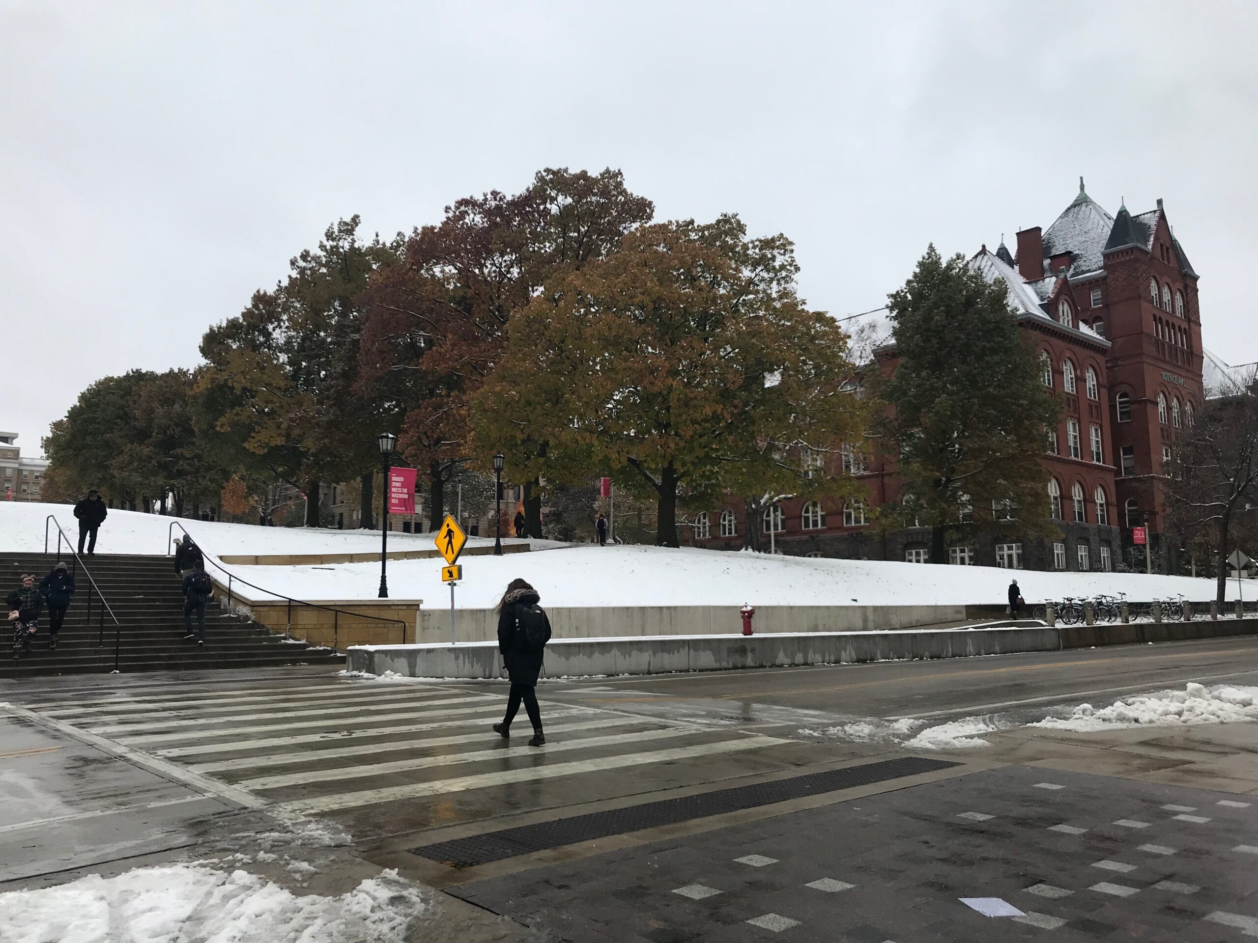 Students cross the street to and from a snowy quad, where the trees still have colorful autumn leaves.