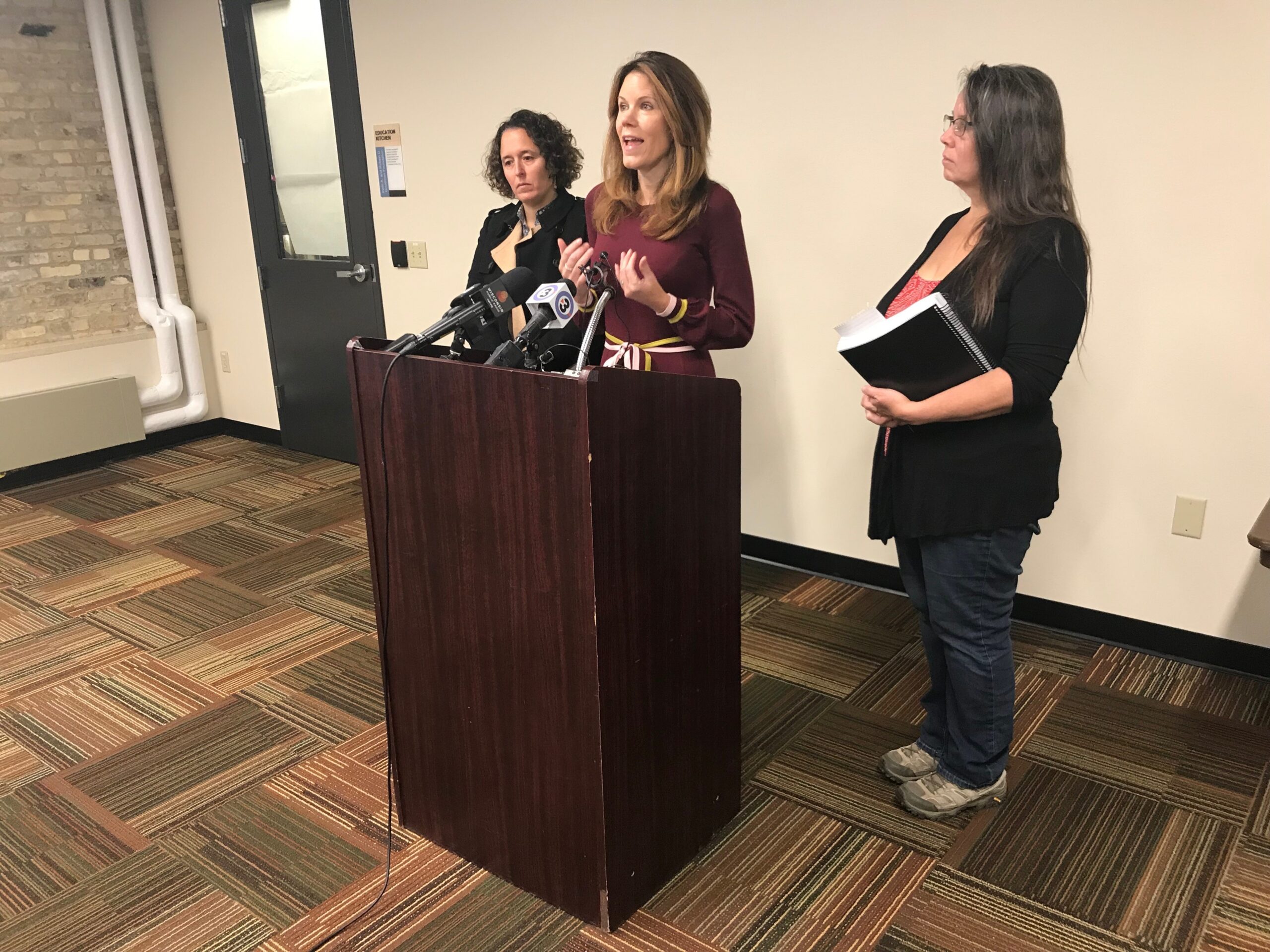 Chris Taylor speaks animatedly at a podium while Amelia Royko Maurer and Rebecca Kemble look on.