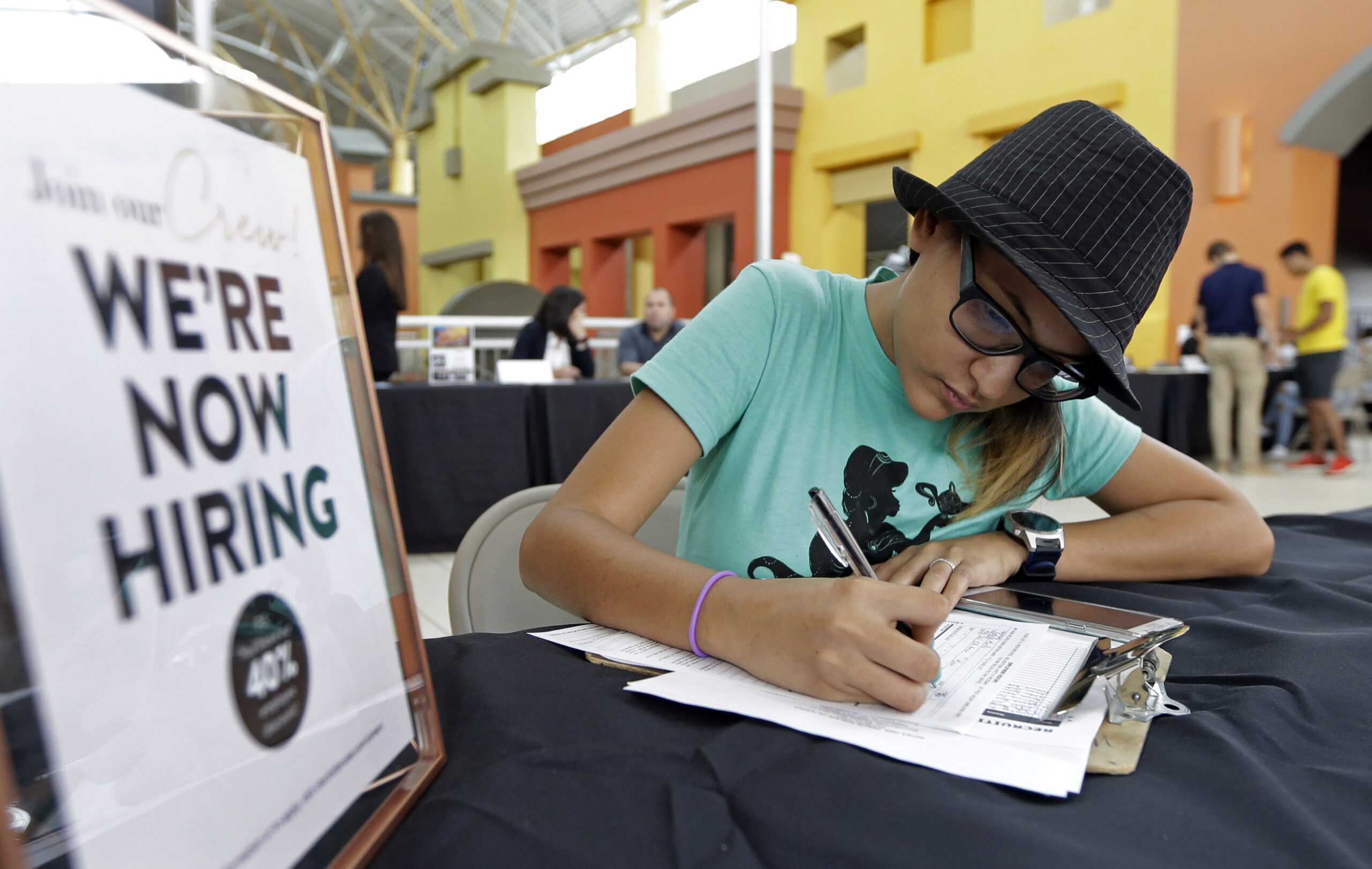 A young woman in a fedora fills out a job application next to a sign that says "We're Now Hiring."
