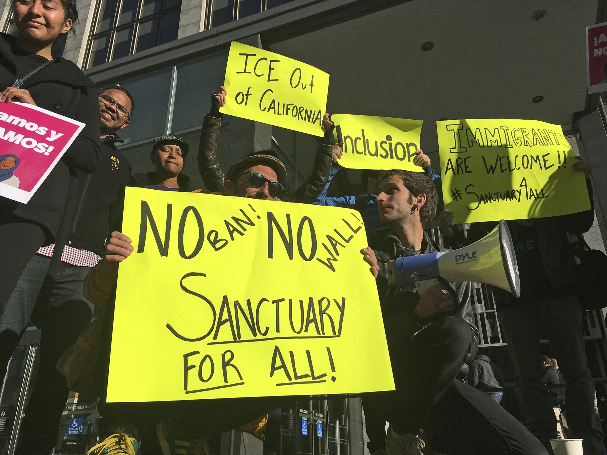 Protesters hold up bright yellow signs reading "No ban! No wall! Sanctuary for all!", "I.C.E. Out of California," and "Inclusion."