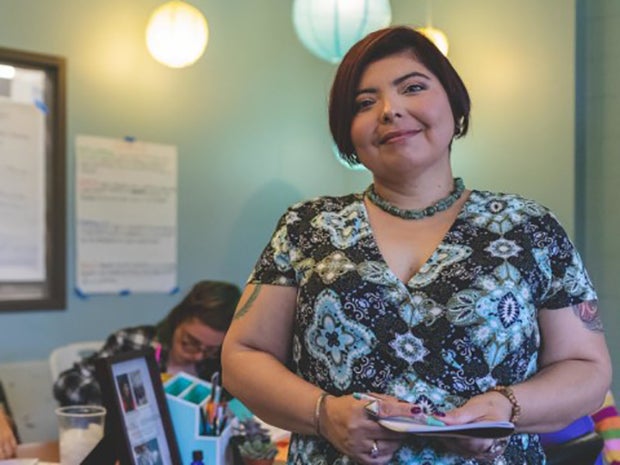 A woman with short hair holds papers in her hands, smiling at the camera in a colorful office.