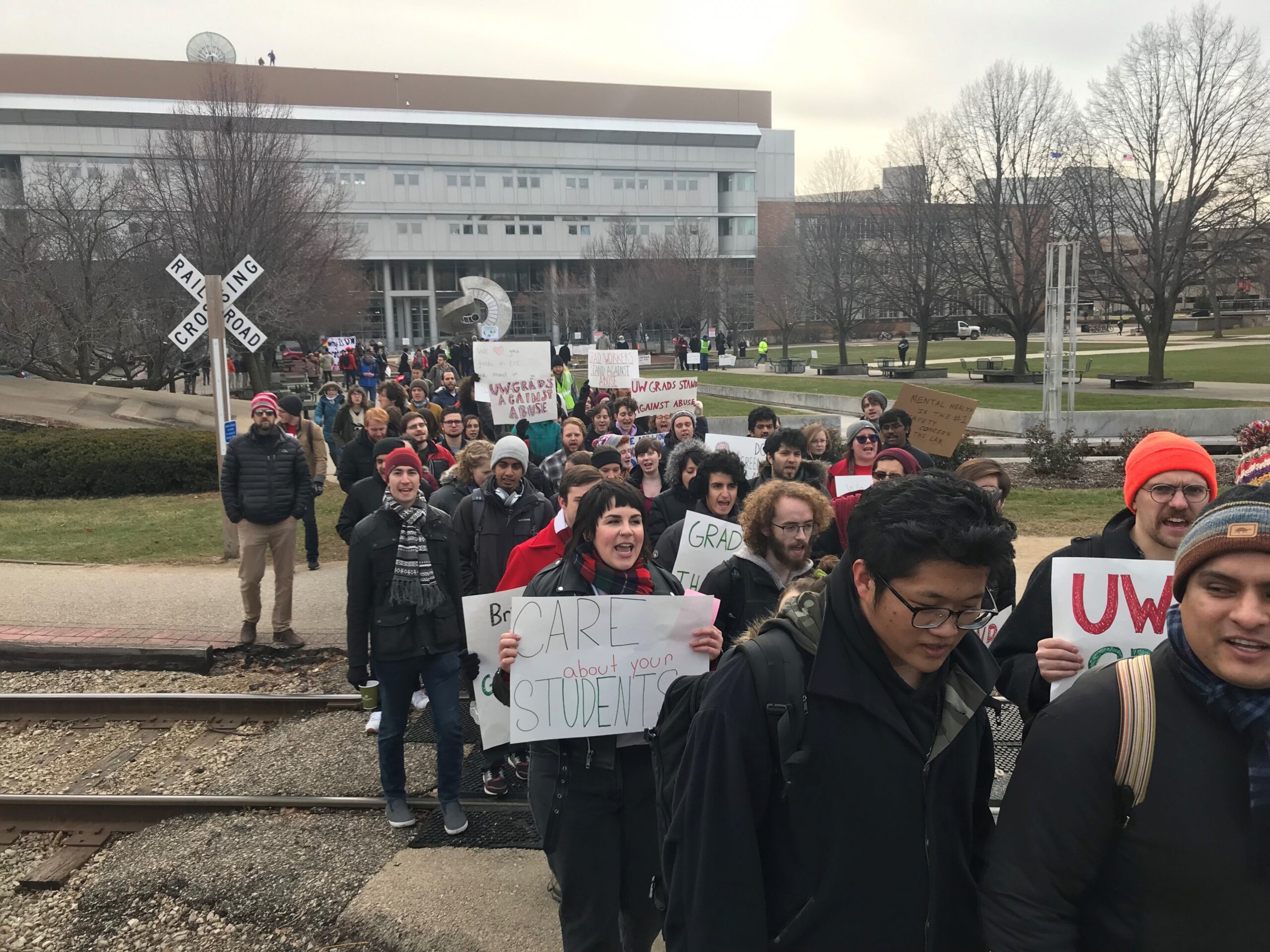 Protestors march across train tracks, holding signs calling for the university to take better care of its students.