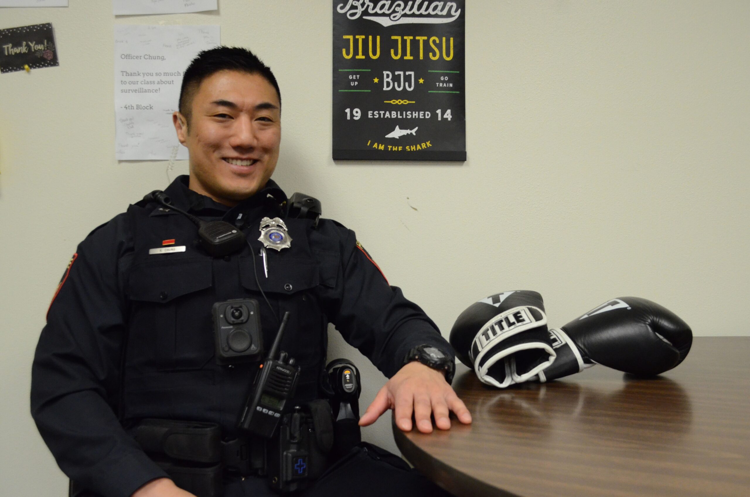 A man in full police uniform sits smiling at a table next to a pair of boxing gloves.
