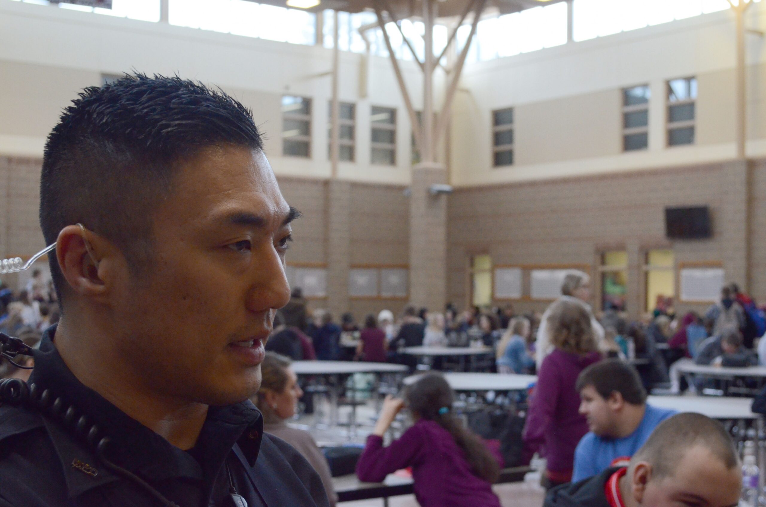 Kenneth Chung, in police uniform with an earpiece, speaks, a full cafeteria of high schoolers sitting behind him.