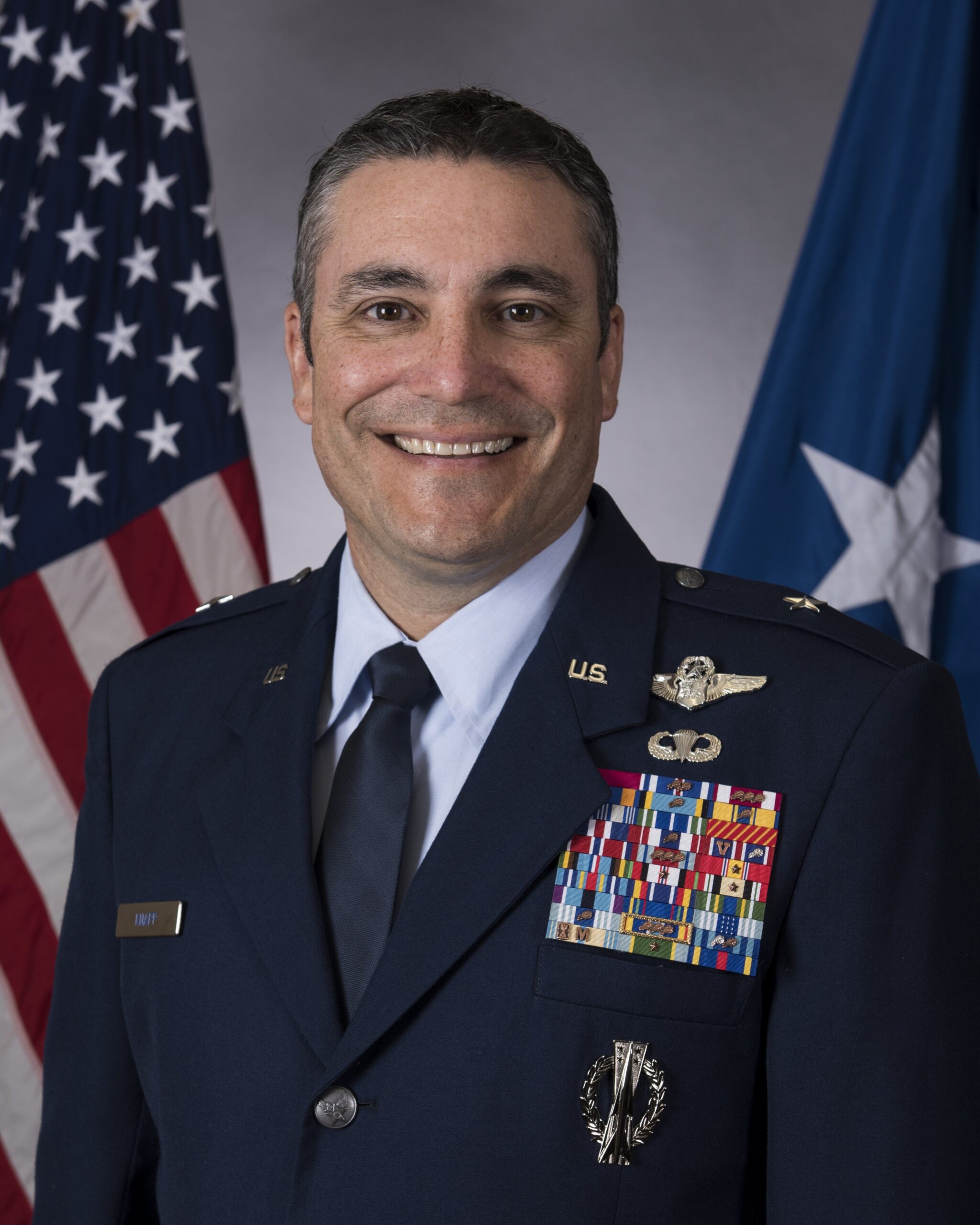 Brigadier General Paul Knapp sits for a portrait in his blue Air Force uniform. His jacket is covered in awards. He smiles in front of an American flag.