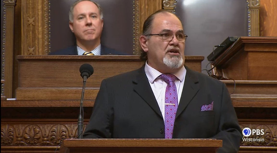 Ned Daniels Jr., a bearded man with a ponytail, stands before a podium, speaking.
