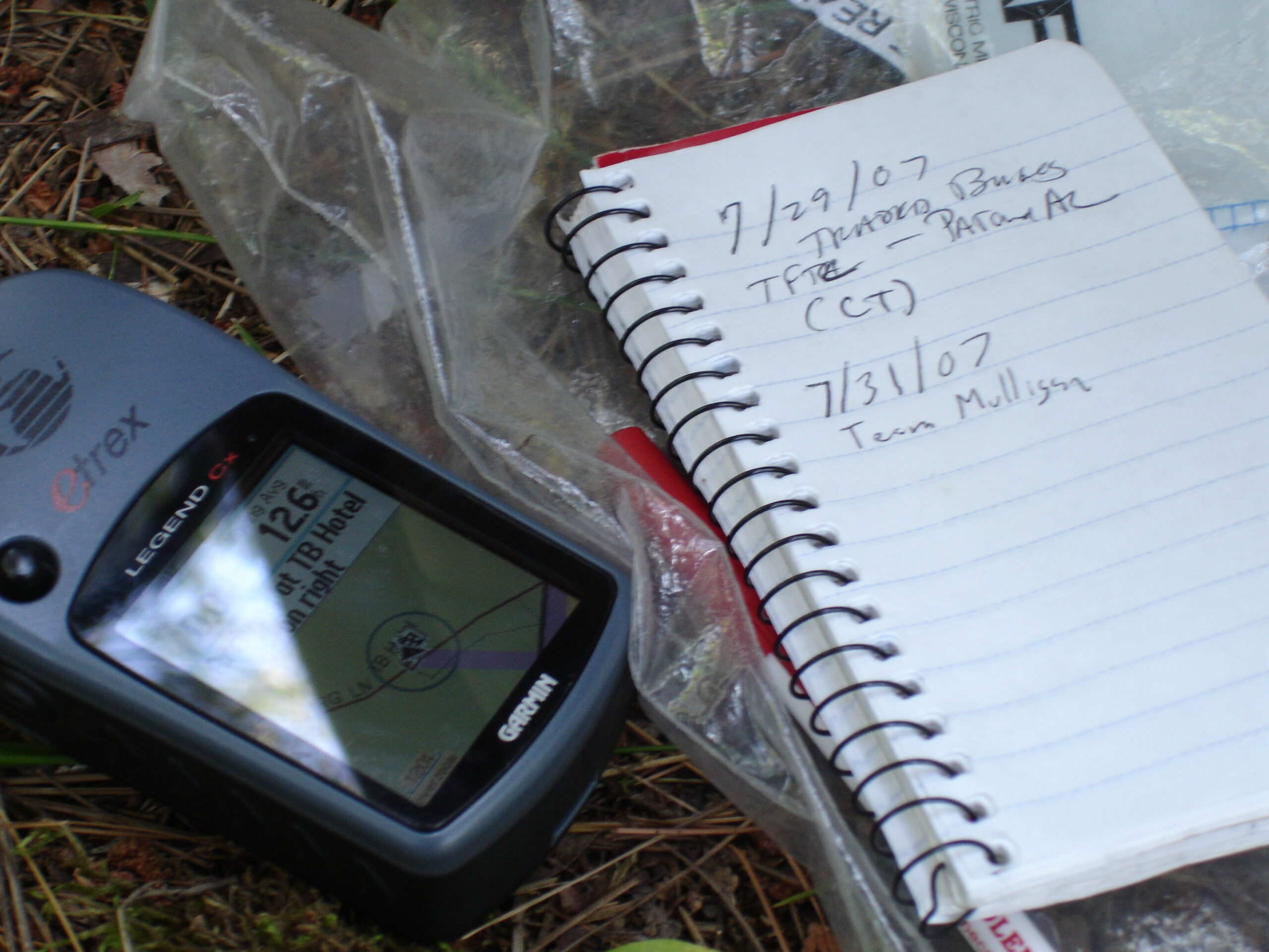 A gps sits on the grass next to the small notebook used as a log in a Wisconsin geocache.
