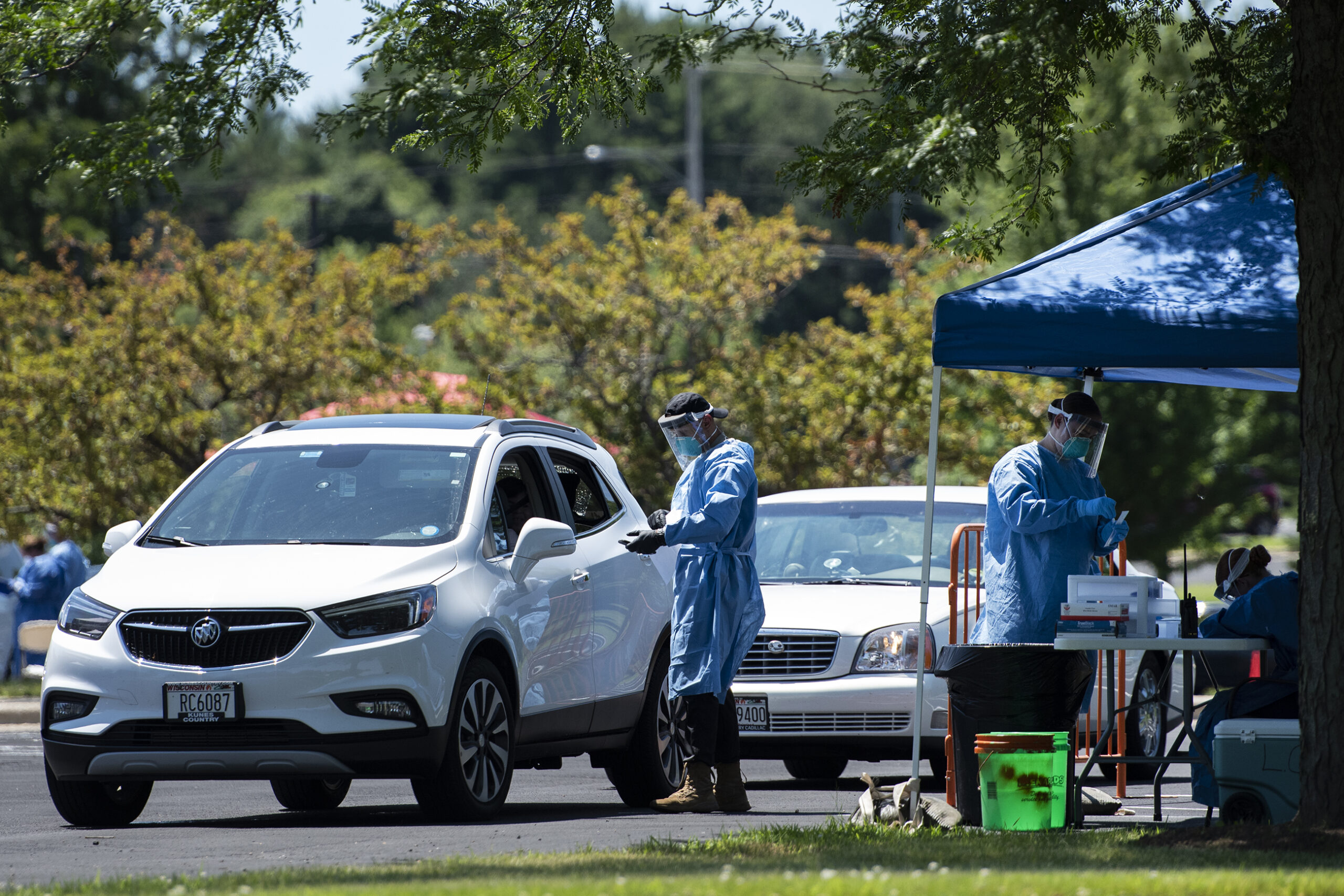 A man in a blue medical gown approaches a white SUV to administer a test