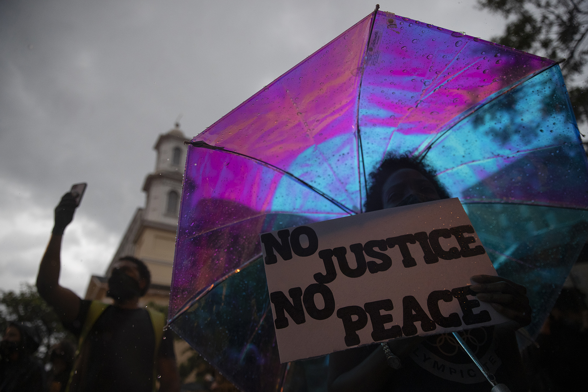 A woman holds a sign that reads "No Justice No Peace" under an umbrella i