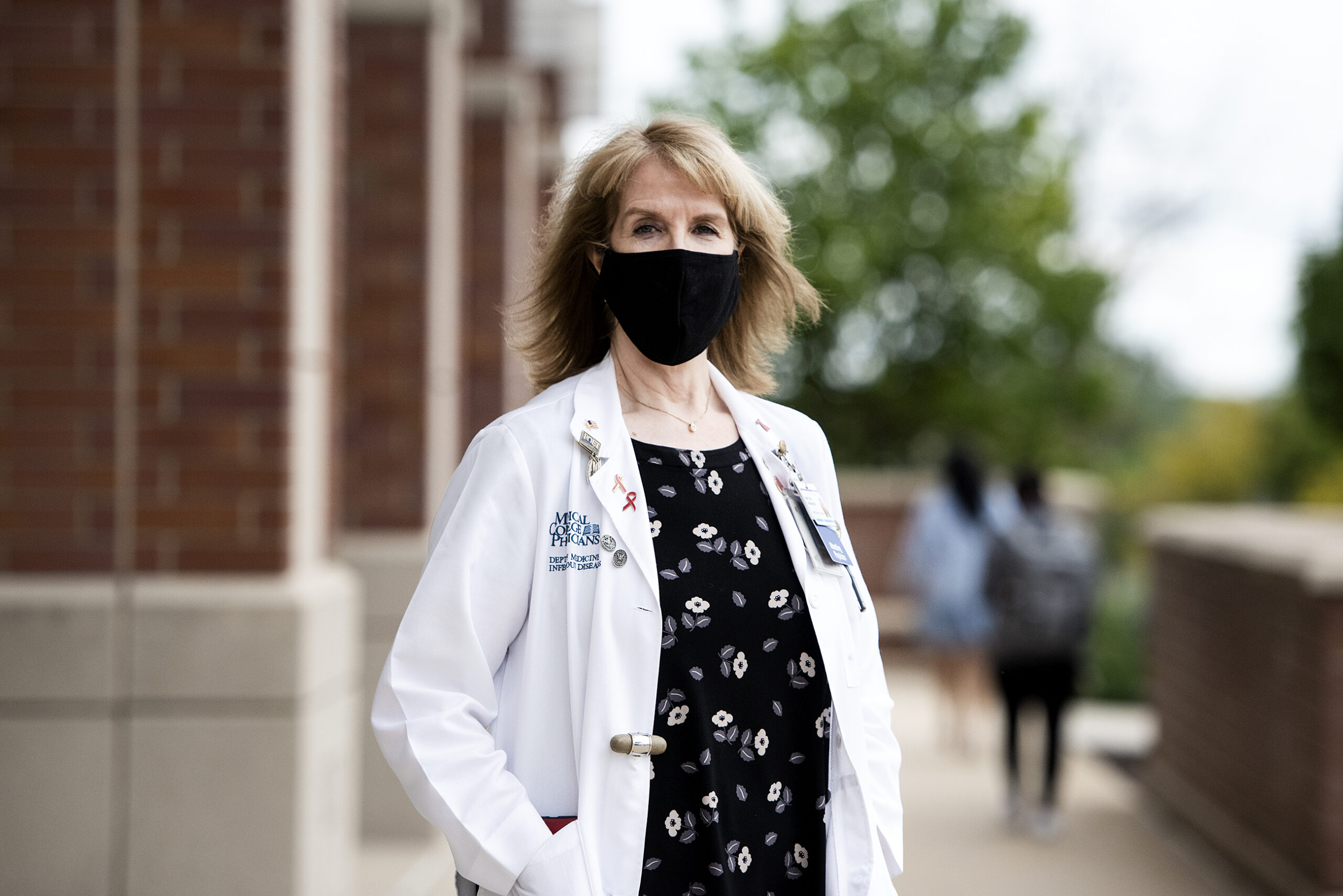 A woman in a white coat and black face mask stands outside as people walk past