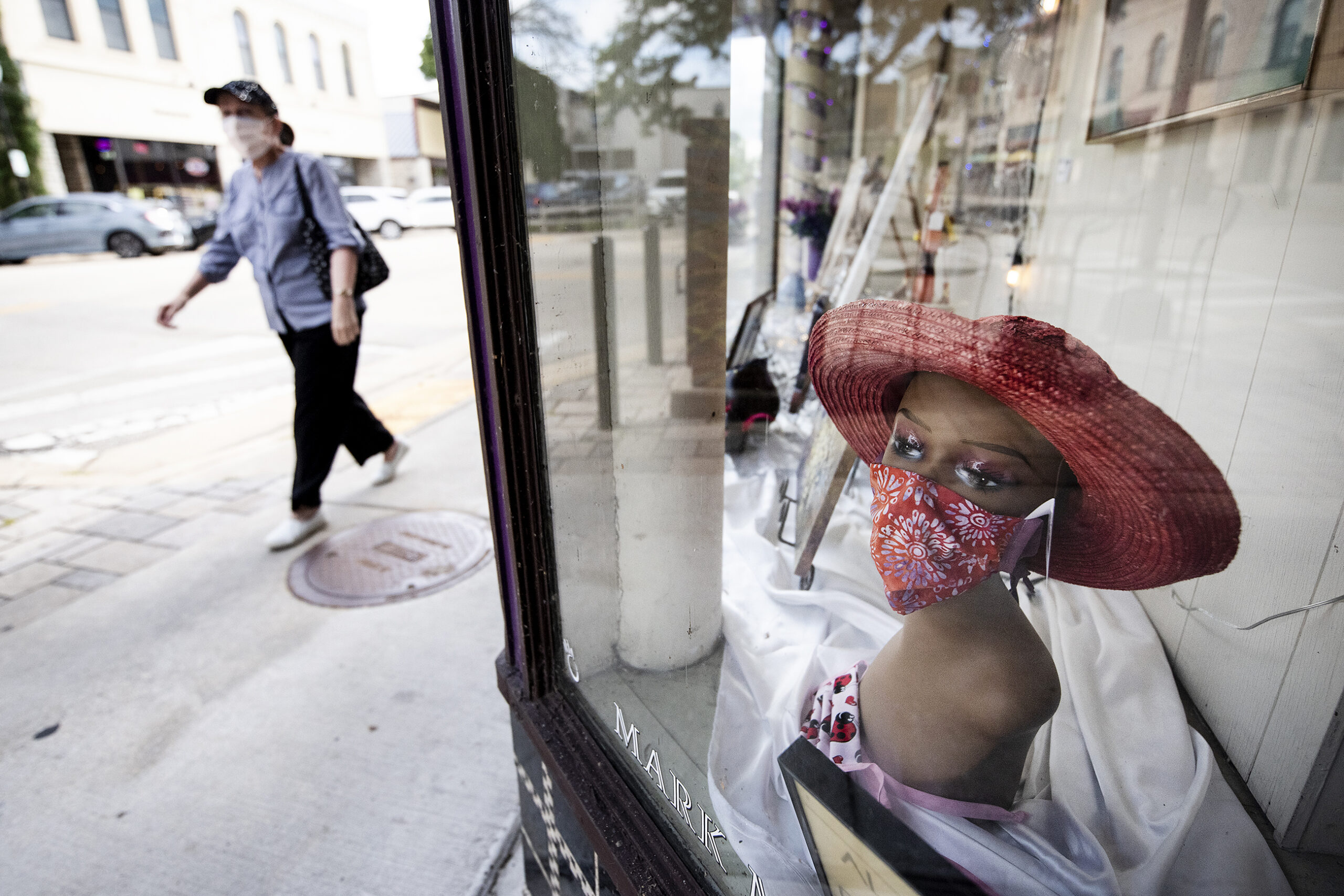 a mannequin bust is used to display a red cloth face mask in a window display as a pedestrian in a mask passes by