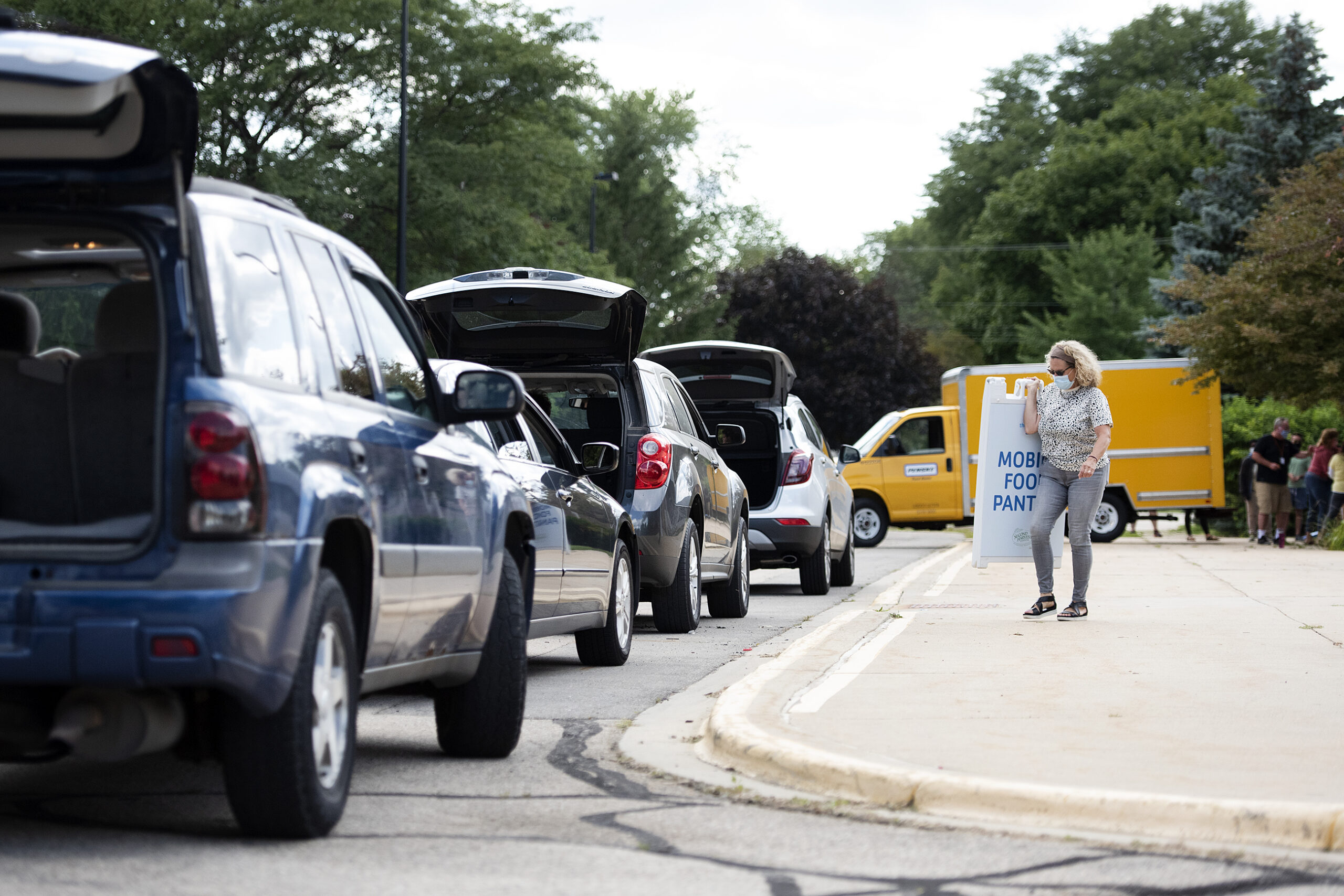 a line of cars with trunks open wait in front of the school as a volunteer walks by with a sign