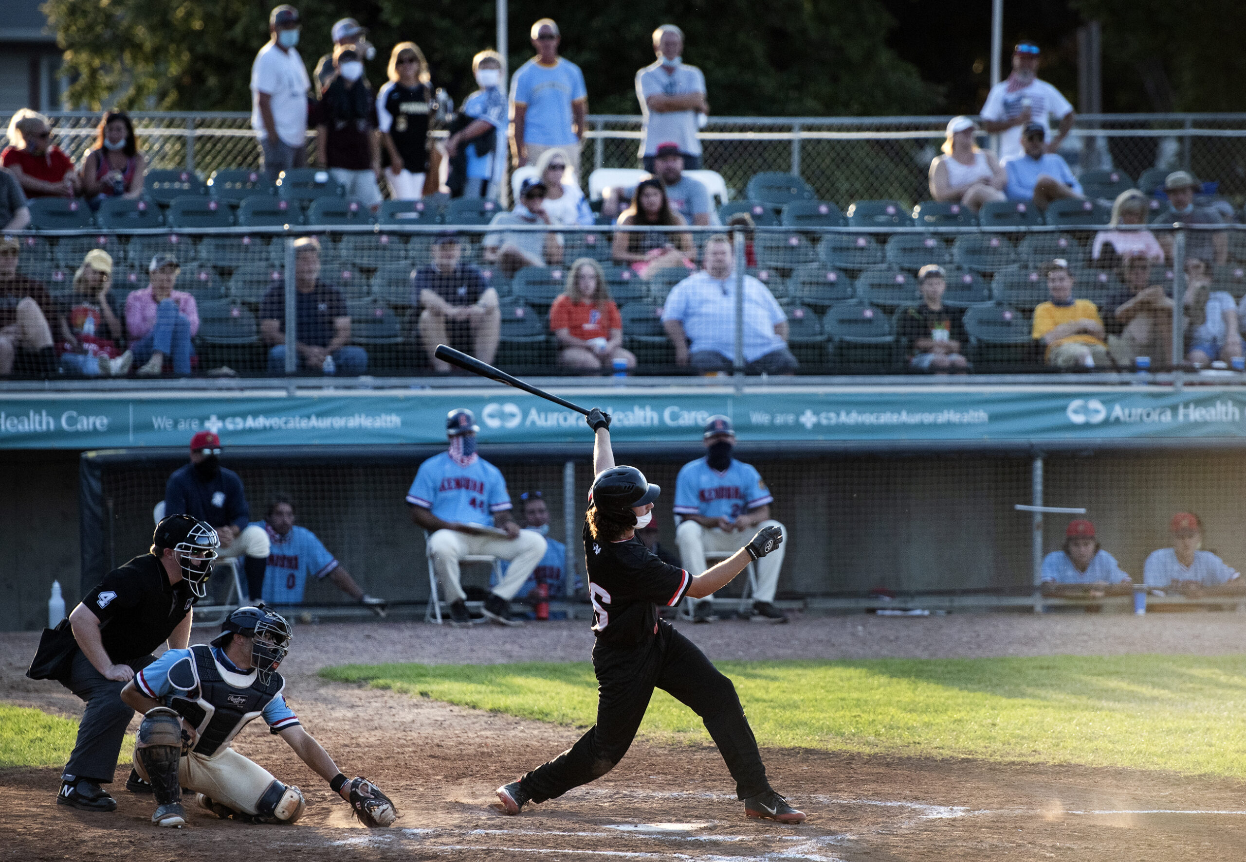 a baseball player in a mask takes a swing as people watch from the stands