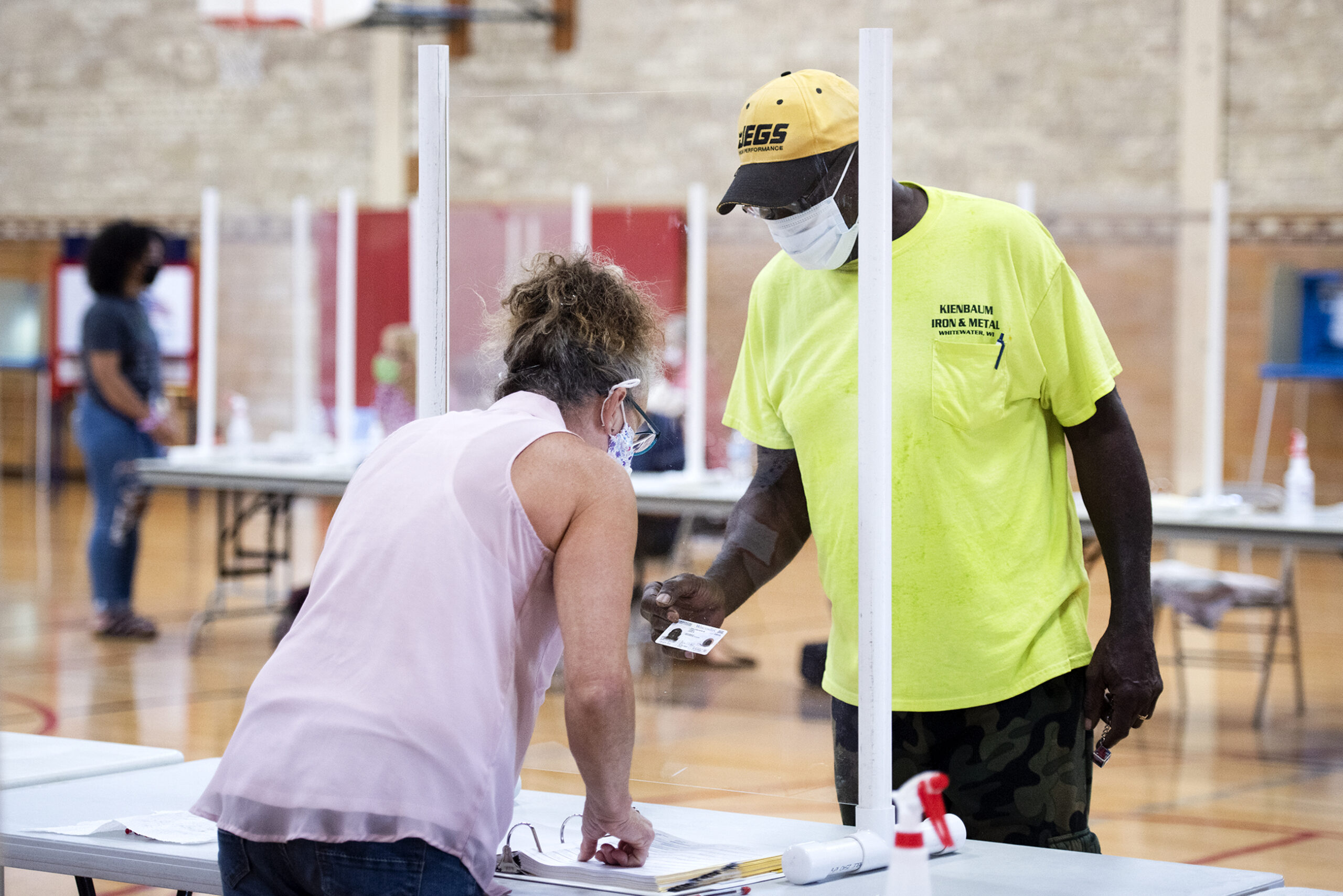 A man in a mask shows his ID as a poll worker views it through a plexiglass barrier