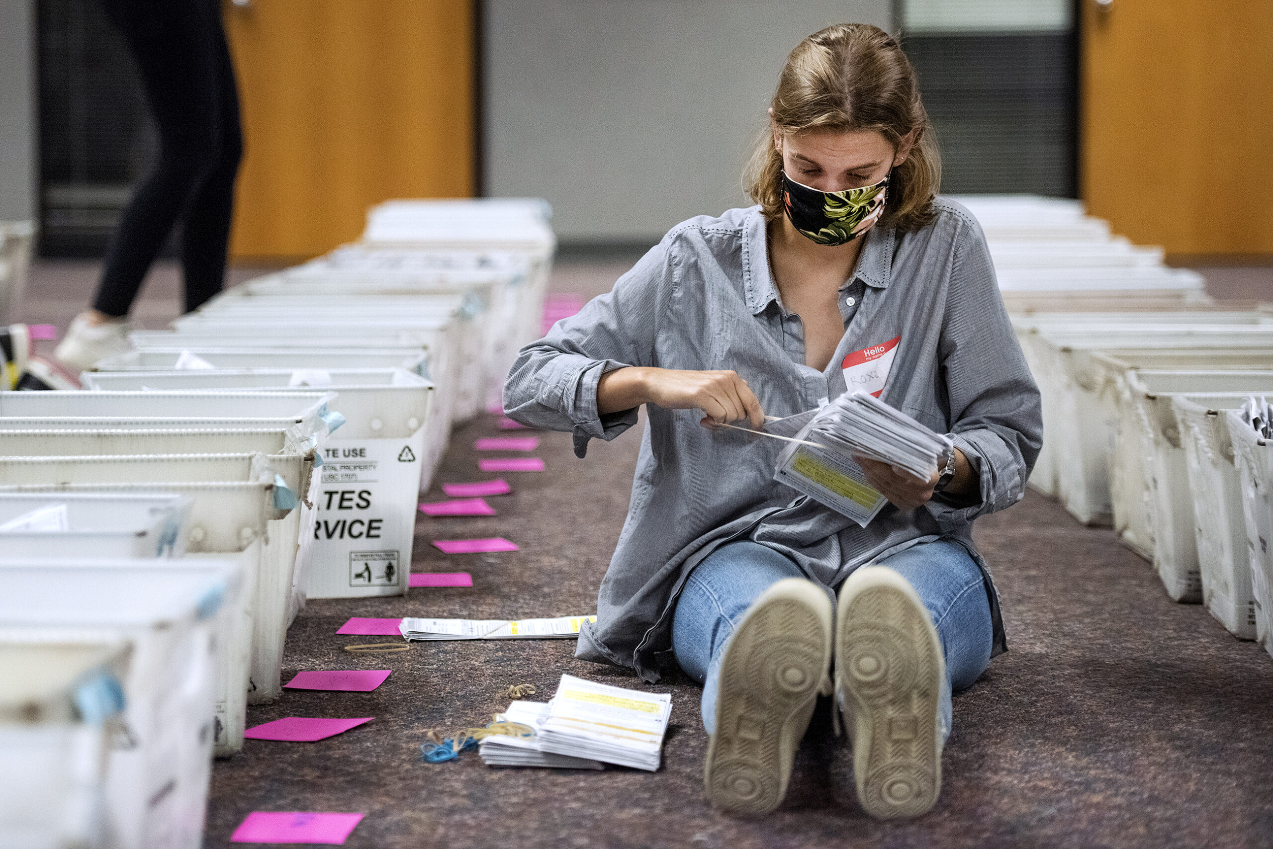 a woman sits on the floor next to rows of bins holding a large stack of ballots