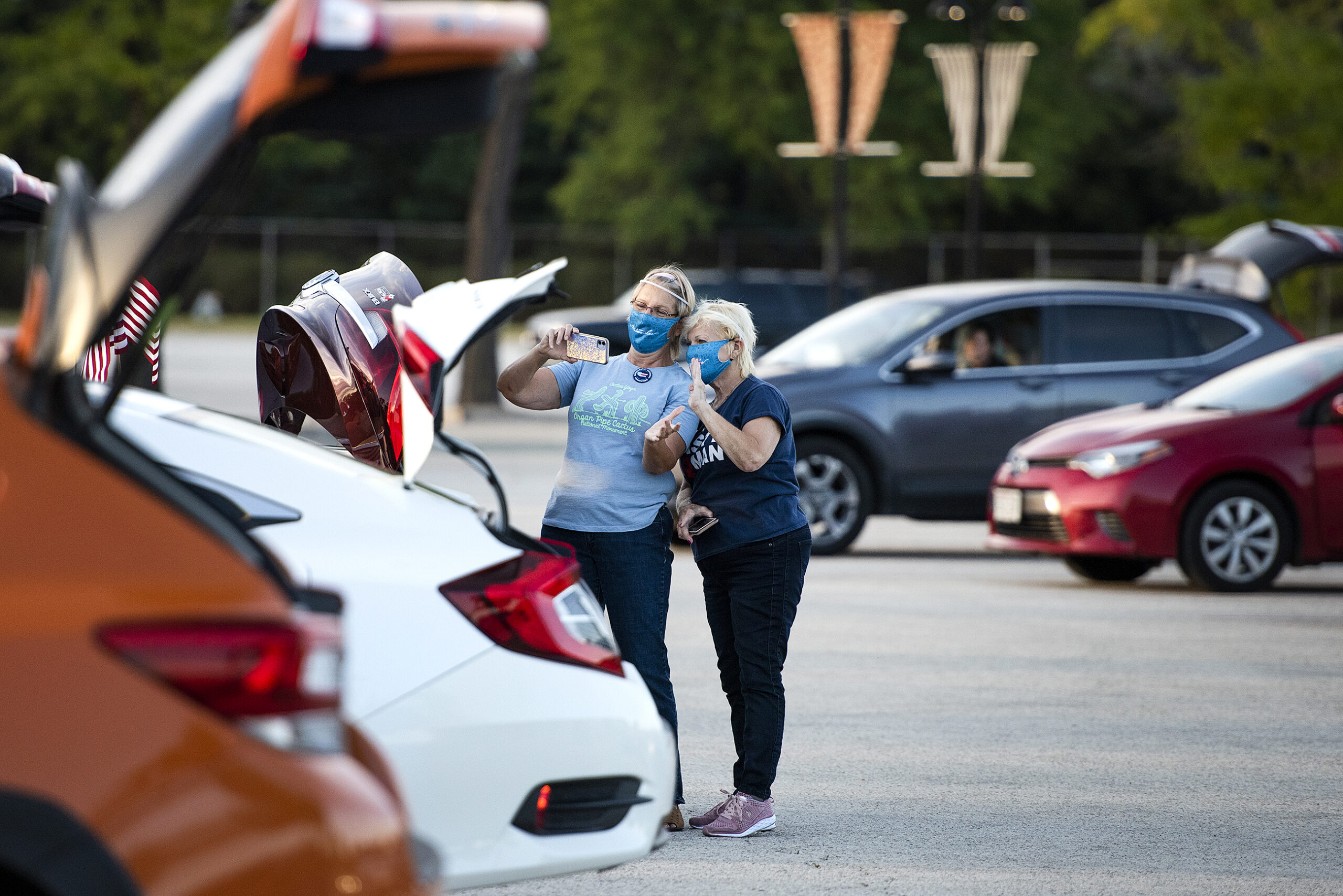 Two ladies in face masks stand near parked cars as they take a selfie together