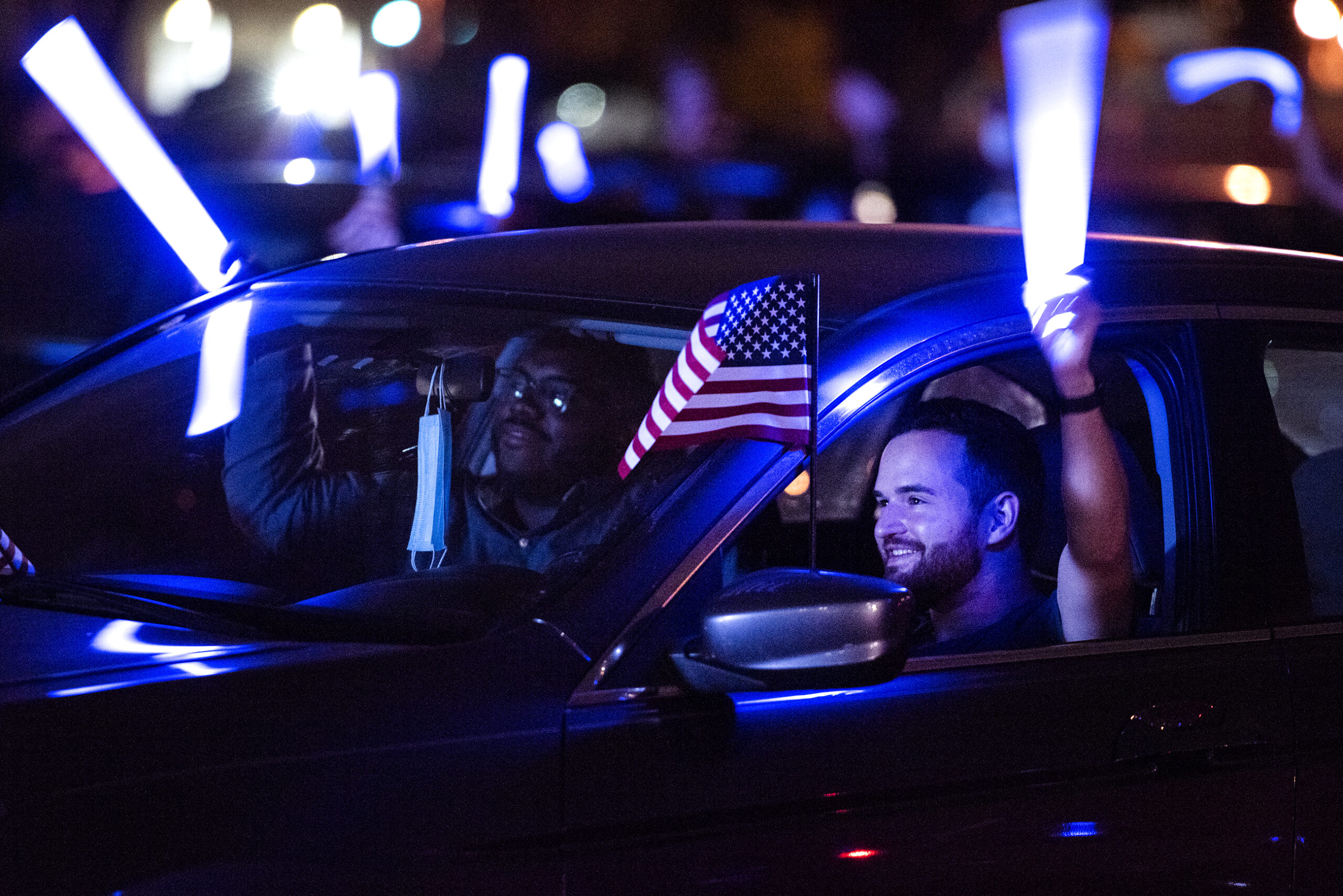 Blue light from a light stick illuminates a smiling man holding an American flag inside his car