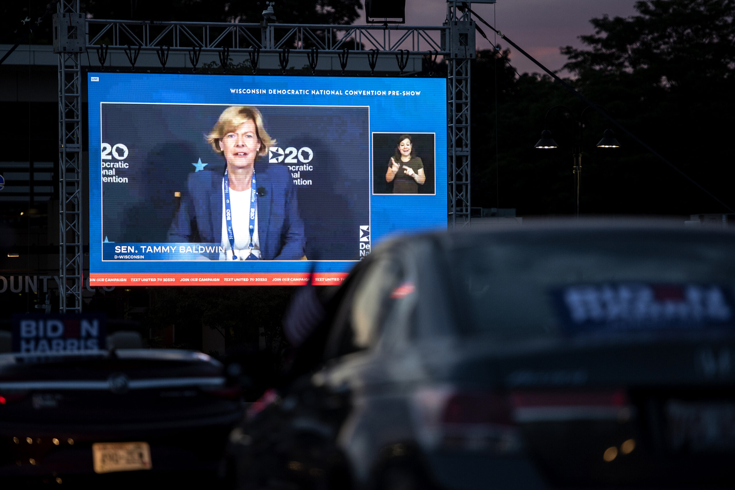 Cars are parked in front of a large screen showing Tammy Baldwin