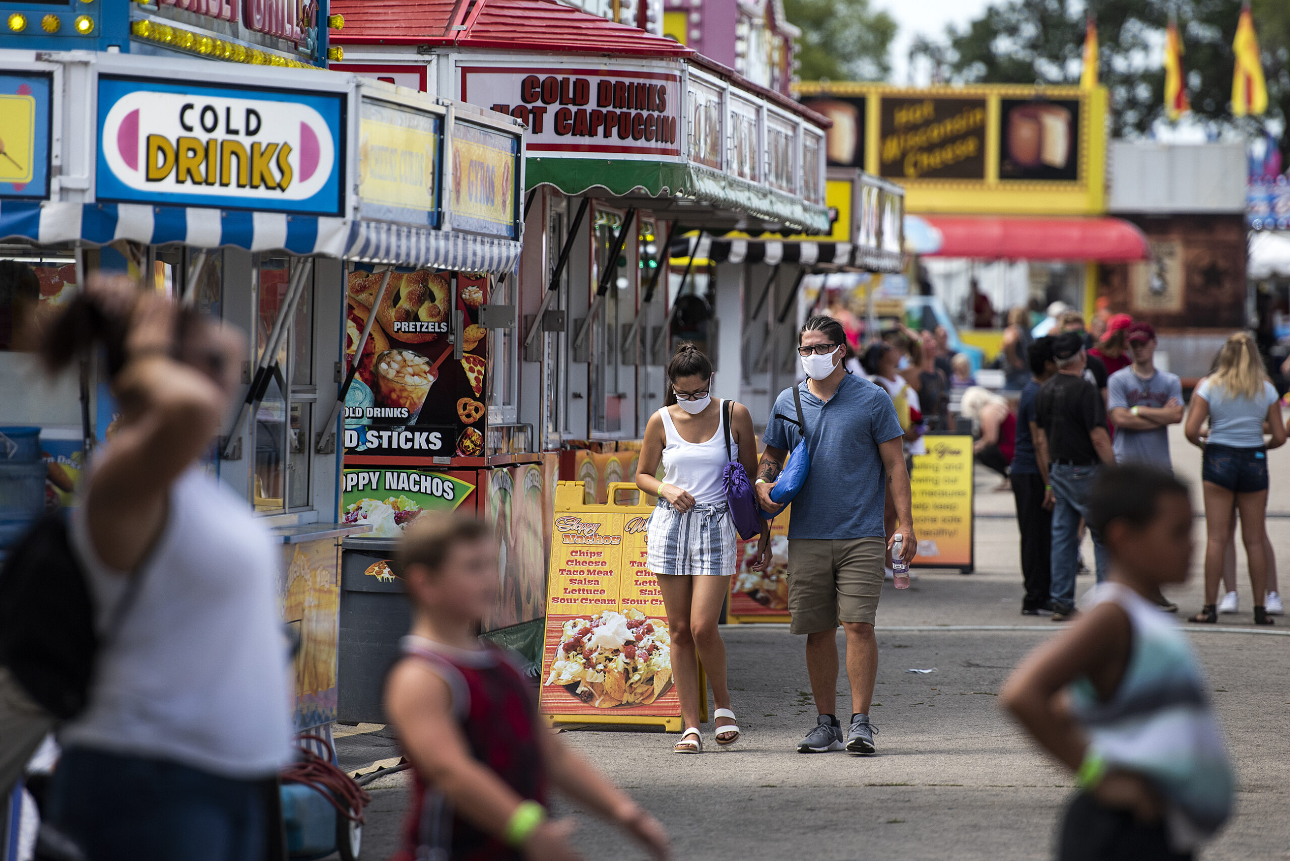 Two people in masks walk in a crowded area of the fair near food vendors