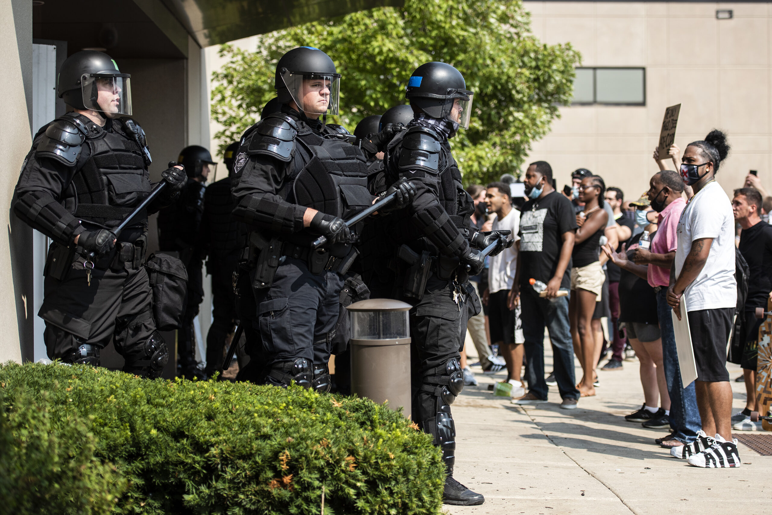 police in riot gear stand with batons next to protesters