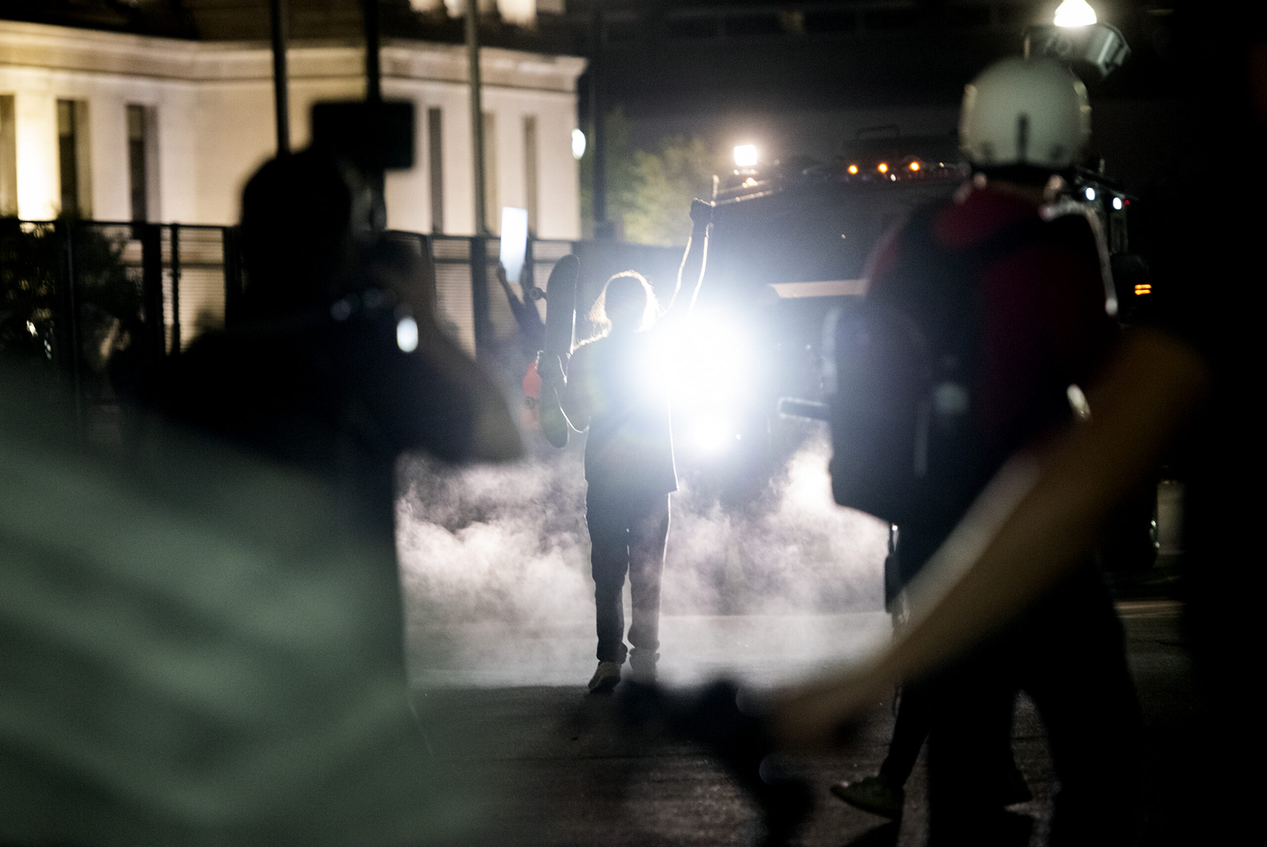smoke surrounds a protester with a fist raised. they are seen in silhouette illuminated by a bearcat headlight.