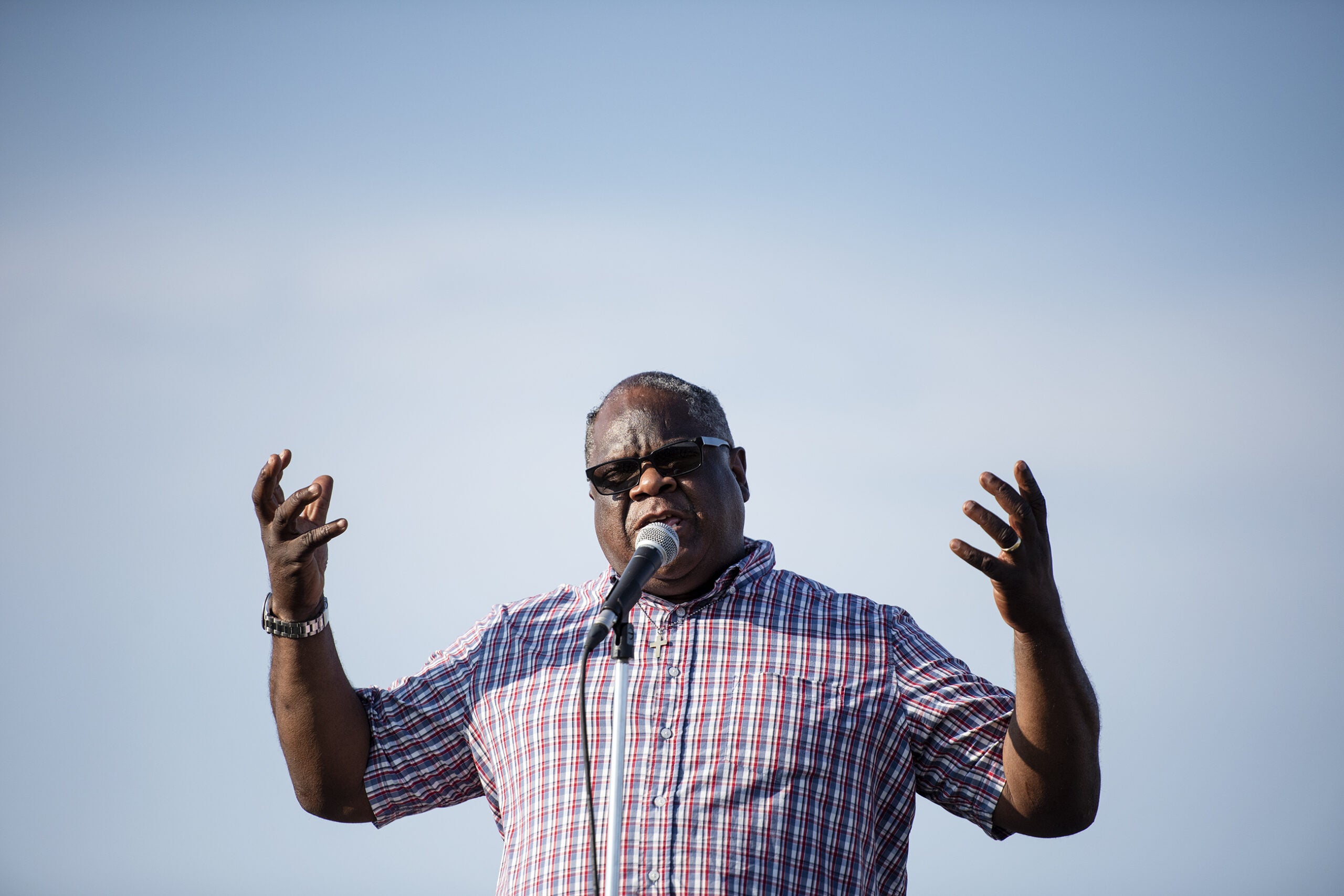 a man shows emotion on his face as he raises his arms and delivers a message backdropped by a blue sky