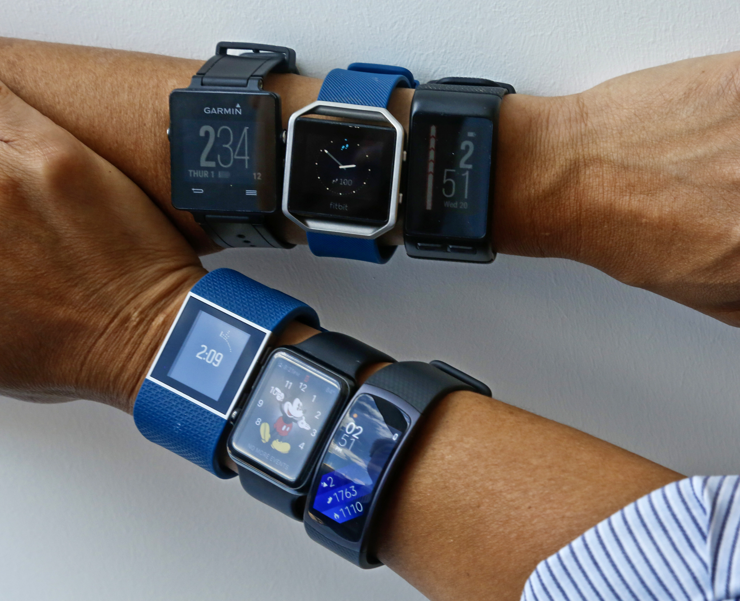 Two wrists display several different kinds of fitness tracker watches with their displays showing the time.