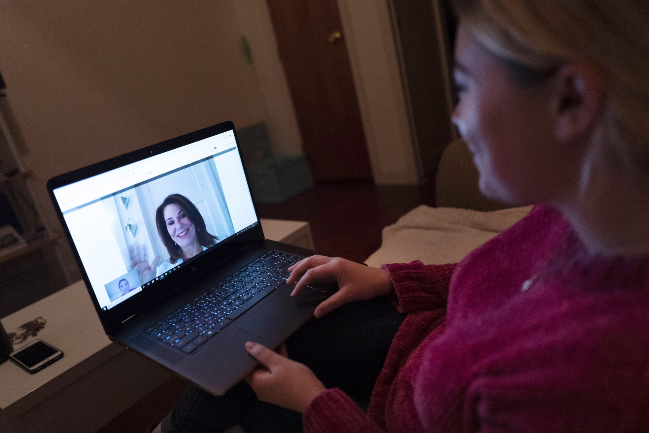 a woman sits on a couch with her laptop screen open talking to her doctor over a video chat.