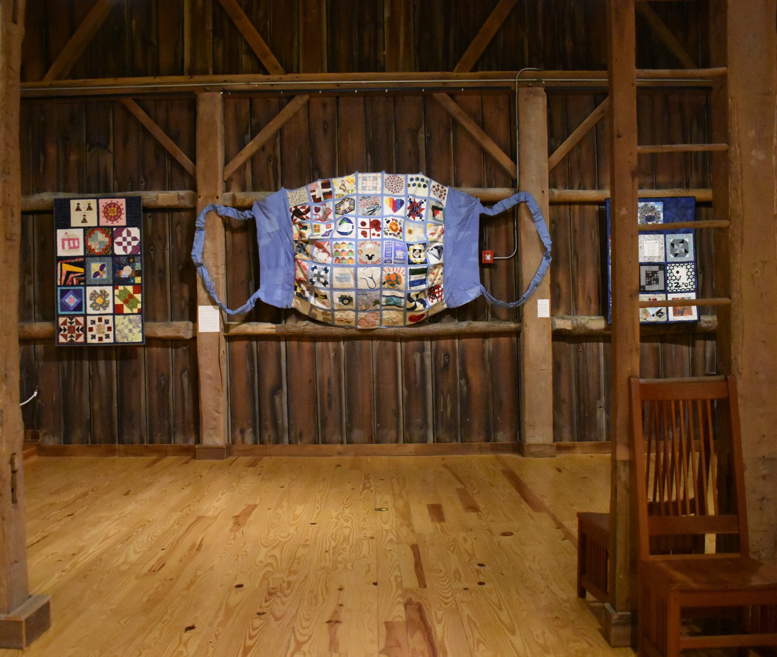A giant mask created out of fabric squares is displayed on the barn-looking wall.
