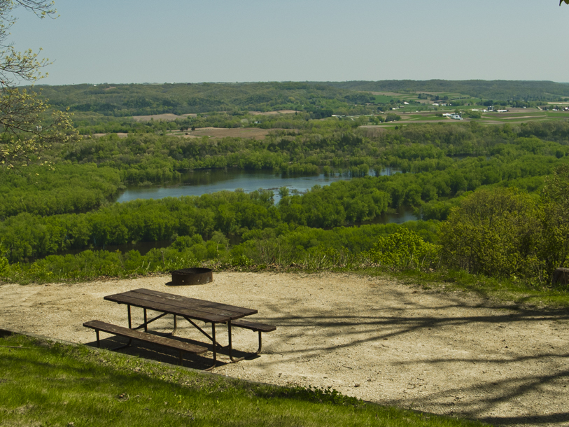A picnic table and firepit of a campsite sit in the foreground with river and green treetops stretching in the background.