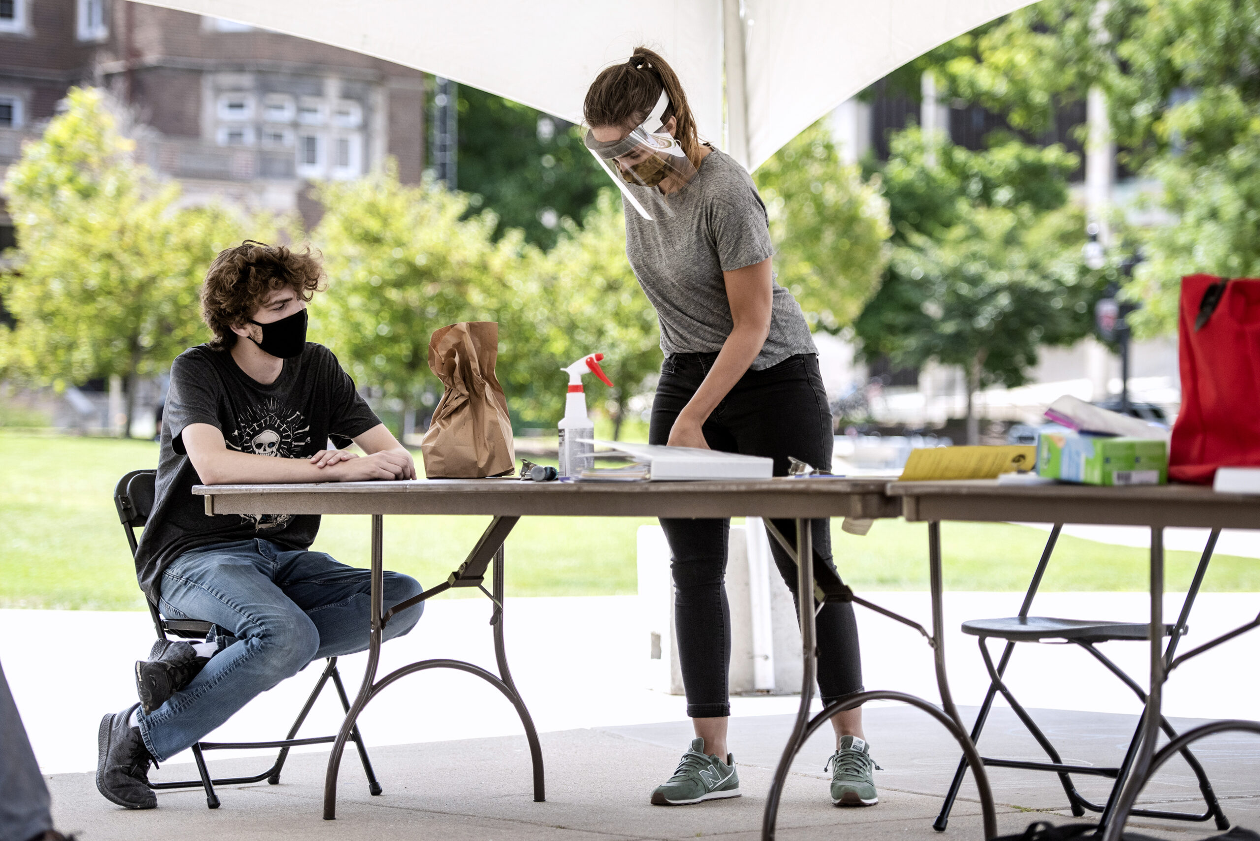 a man in a mask sits outdoors at a table as a poll worker in a face shield gives instructions