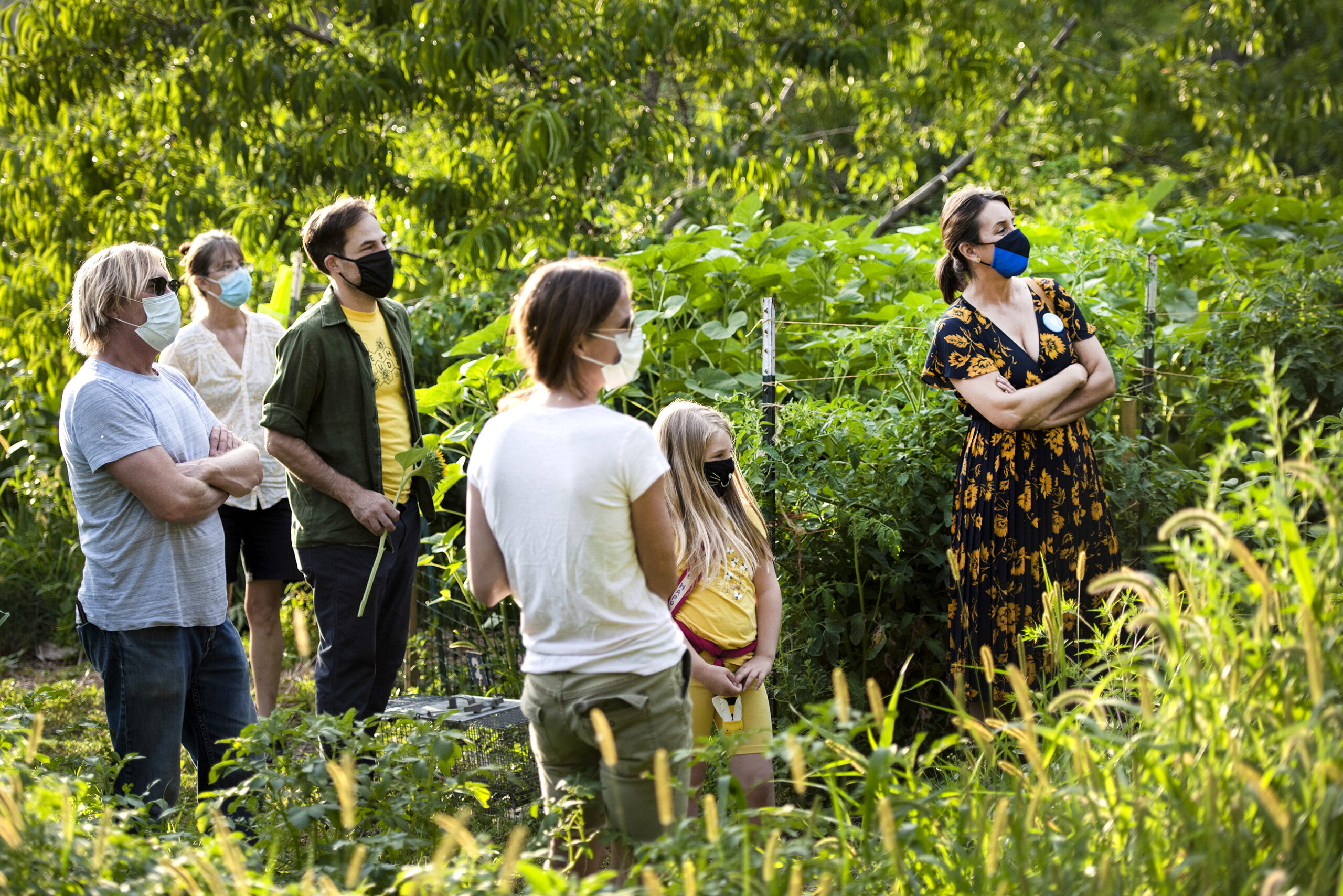 a group of people in face masks are surrounded by greenery as they listen to a speaker
