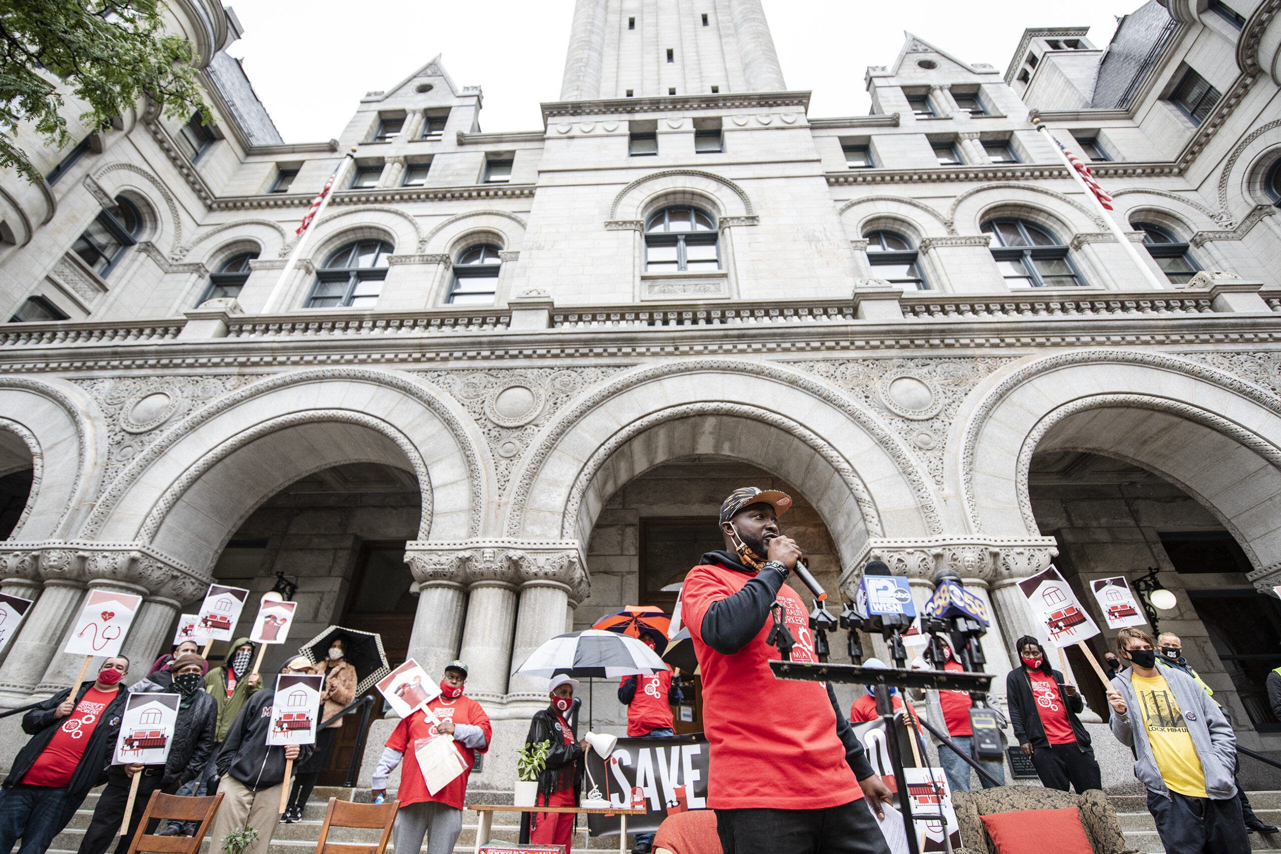 a man speaks in front of a crowd holding umbrellas. a large white building can be seen towering above him.
