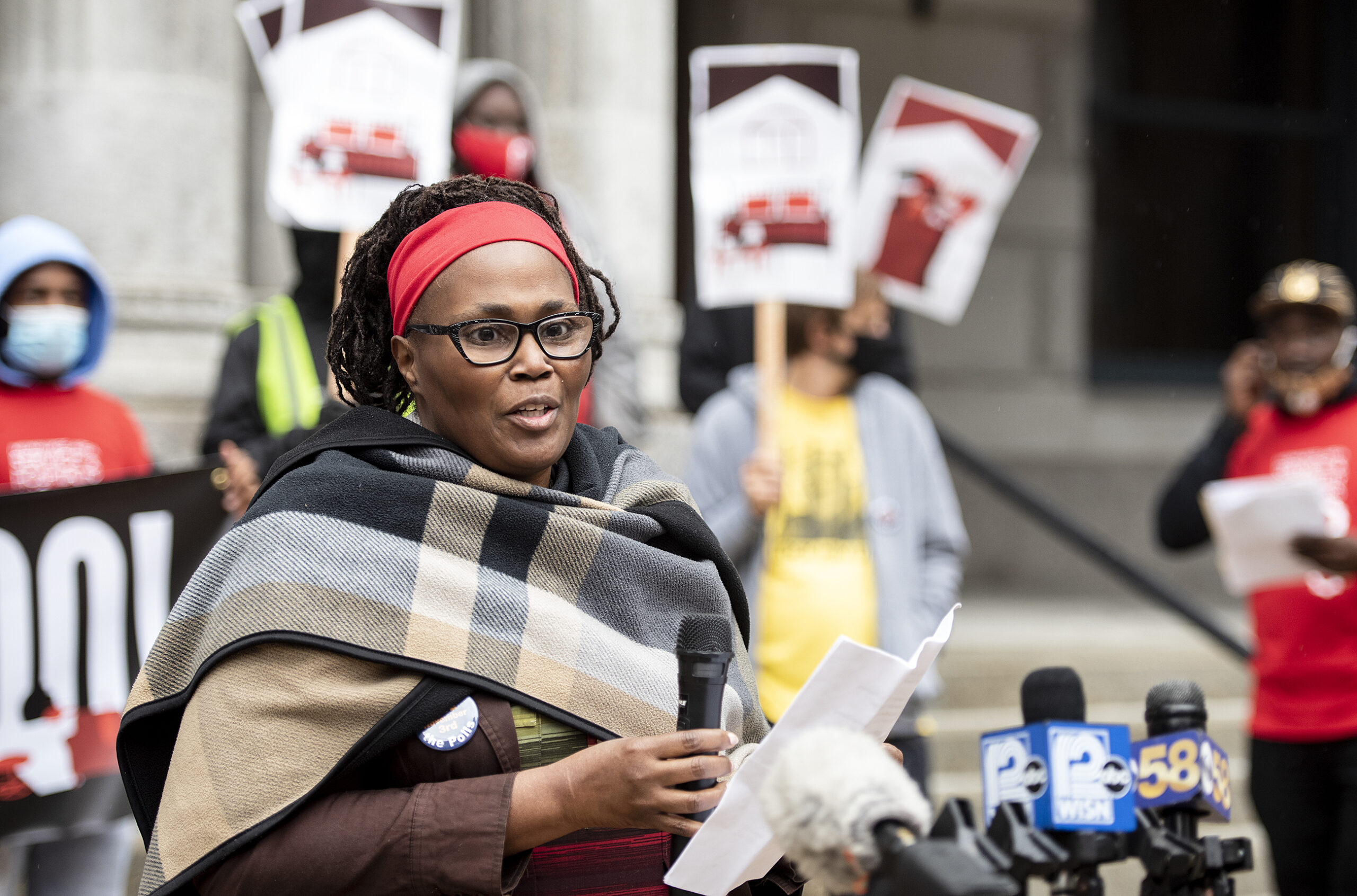 a woman speaks into a microphone as attendees hold signs behind her