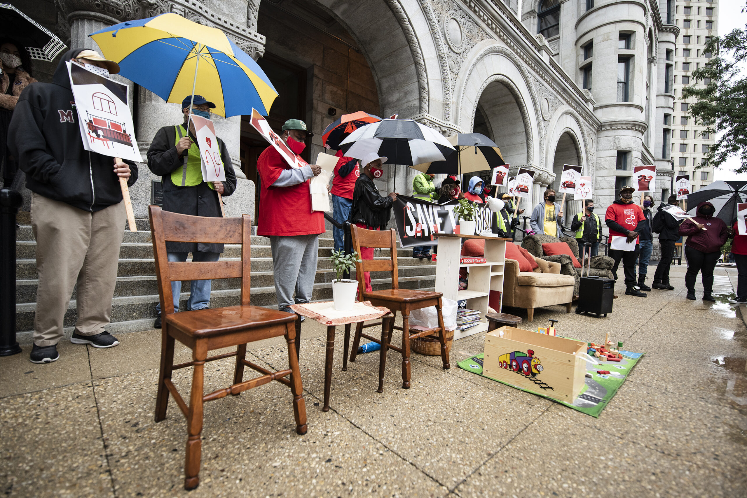 two chairs, a bookshelf, a couch, and some toys on a rug are set up in front of protest attendees