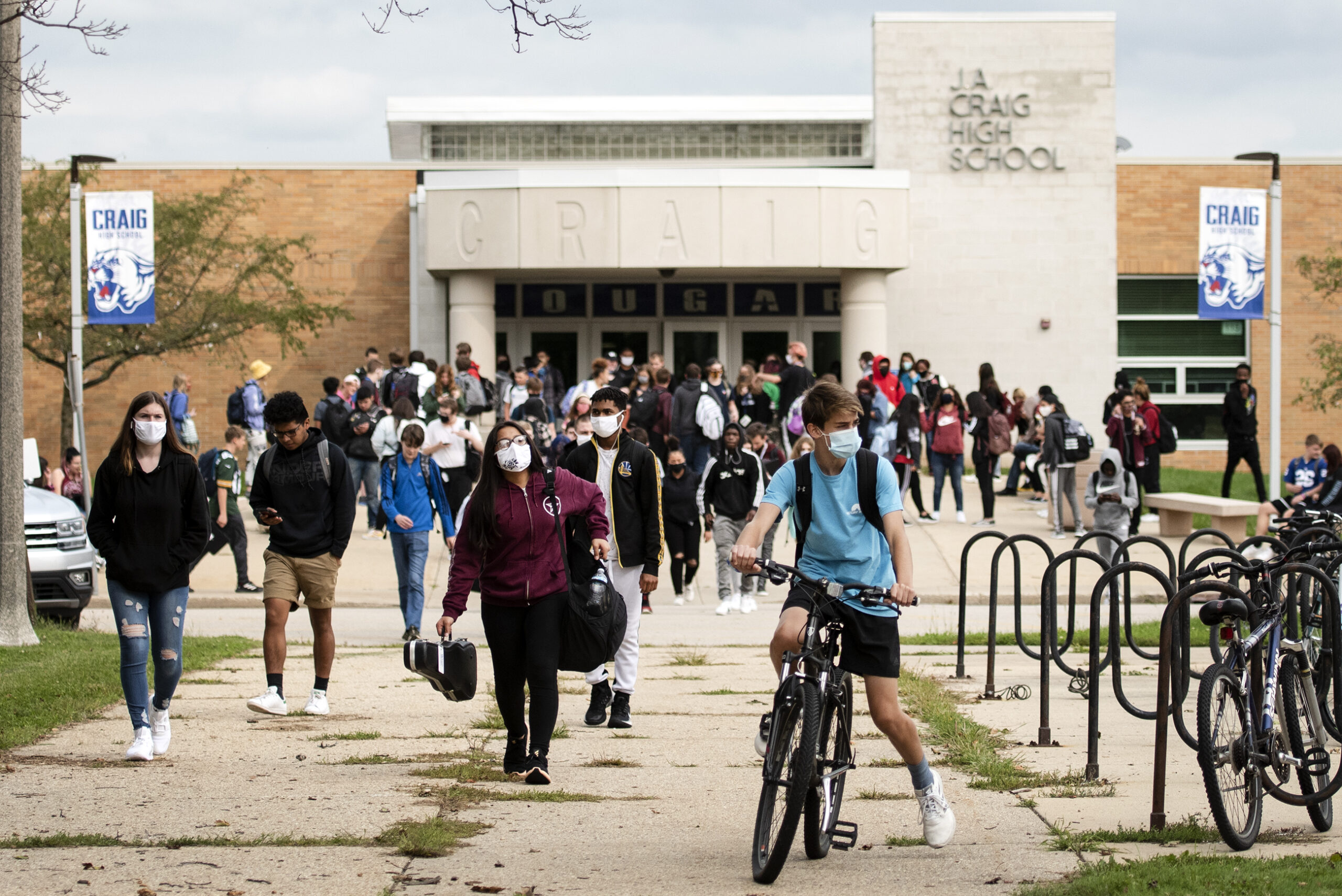 a student in a mask rides a bike as a sea of high school students, some wearing masks, exit the school