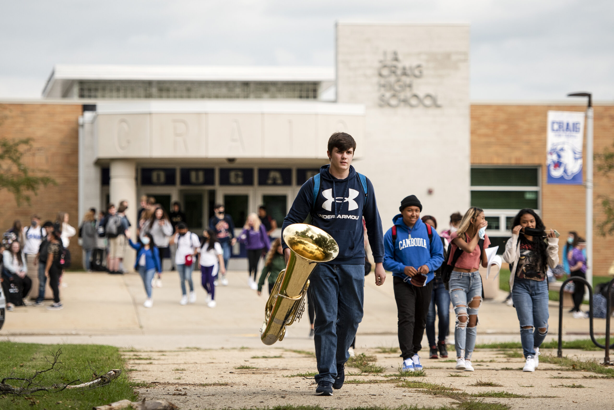 a student carries a tuba in his hand in a crowd of other students exiting a high school