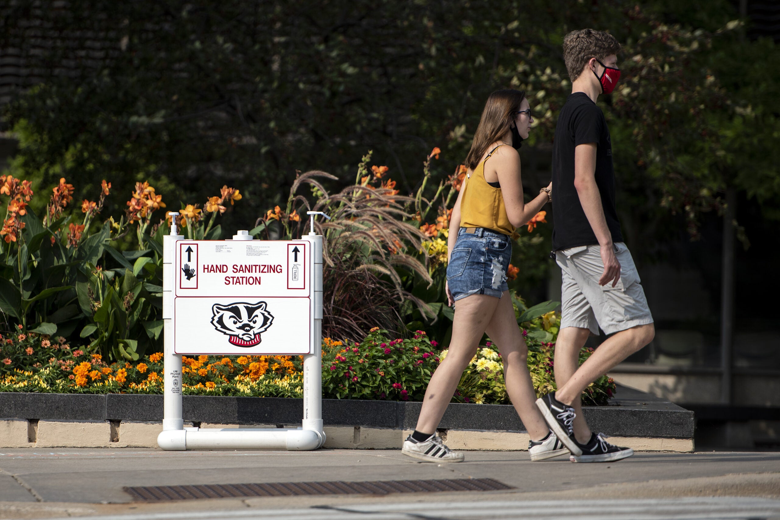 two people walk by some hand sanitizer on a small stand displayed with the image of Bucky badger
