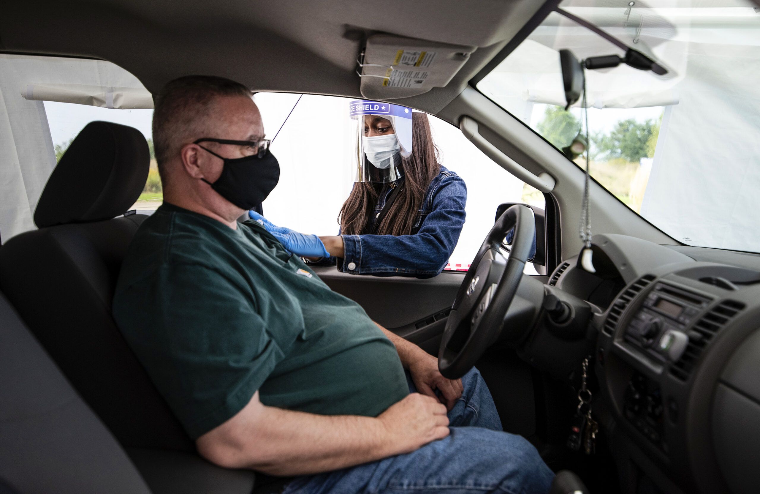 A woman in a mask and face shield reach into a vehicle to give a flu shot to the driver