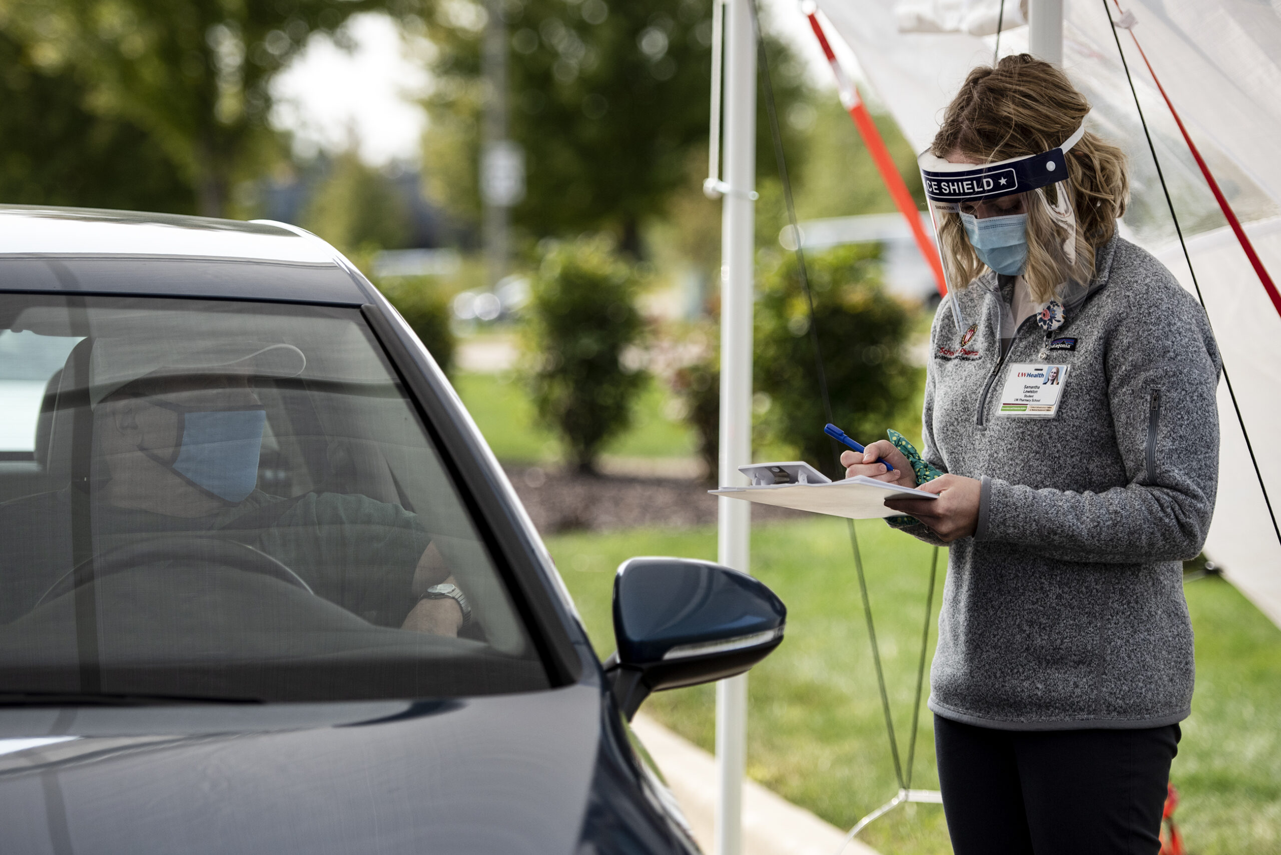 a woman in a mask and face shield writes on a clip board while standing under a white tent outside set up for the drive thru service