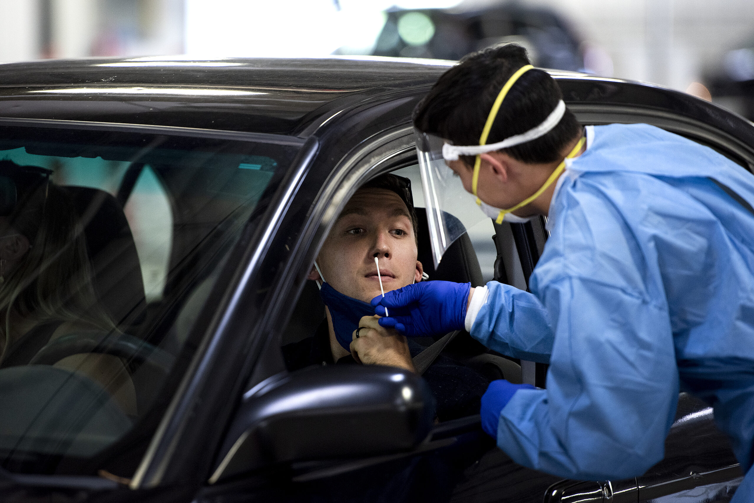a man remains in his car as a white swab is inserted into his nose by a man in a blue medical gown and gloves