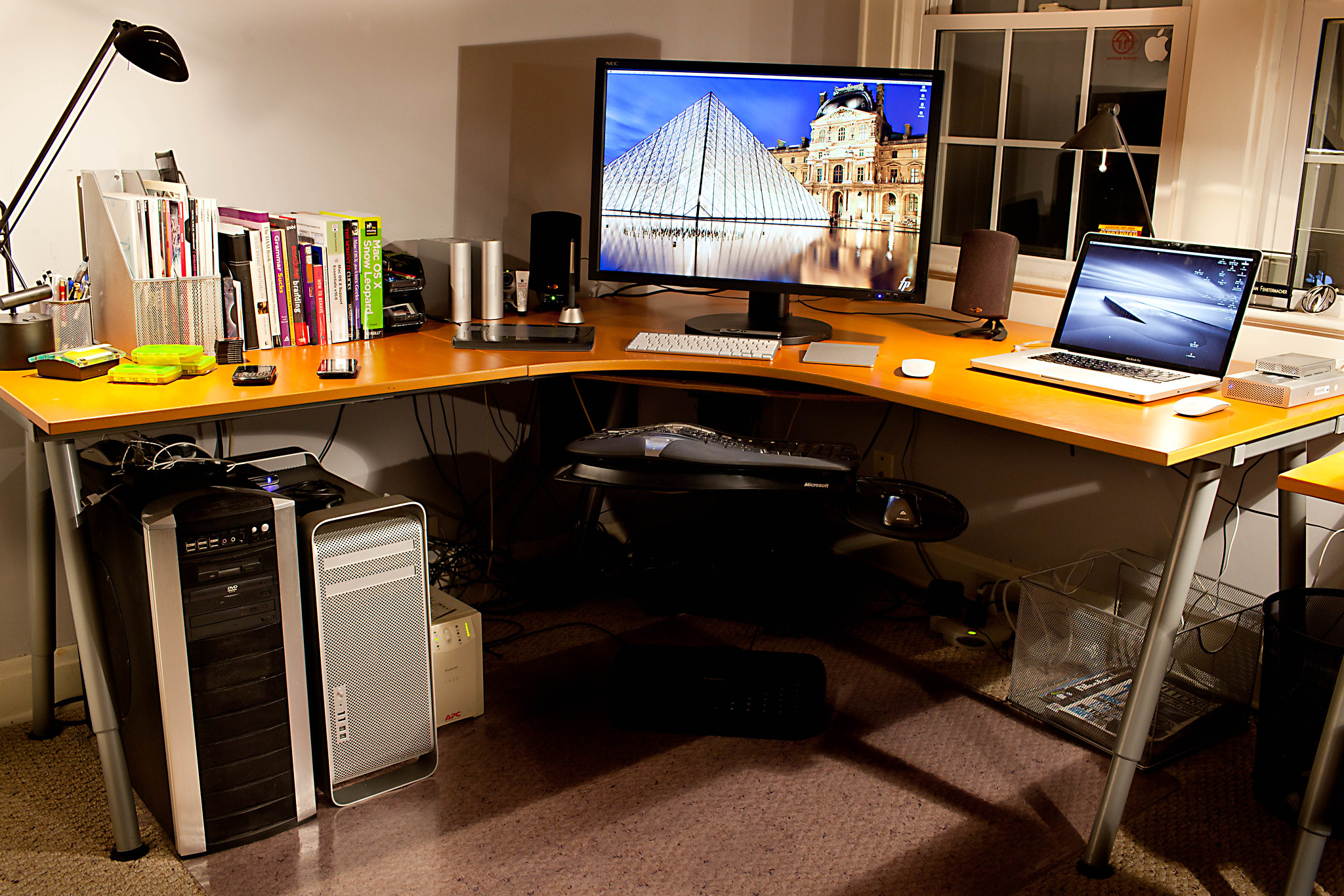 A large corner desk with books, computer screen and laptop on the top and two modems underneath.