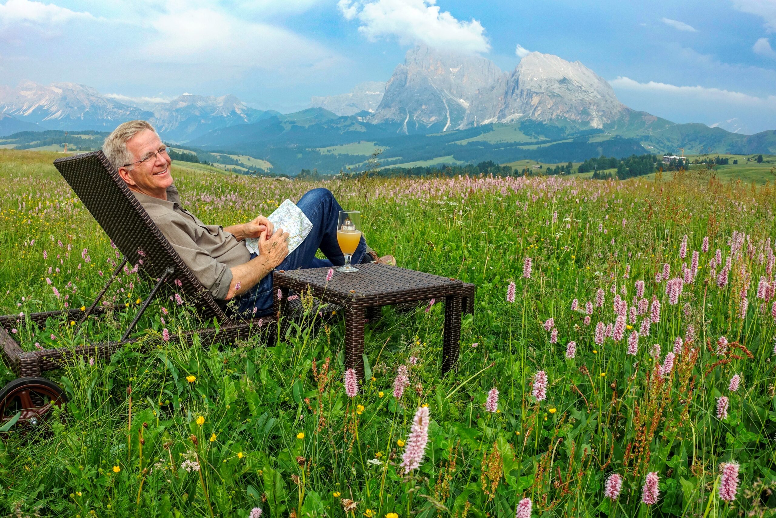 Rick Steves sits in a lounge chair with a map in hand in the middle of a lush green field with mountains in the distance.