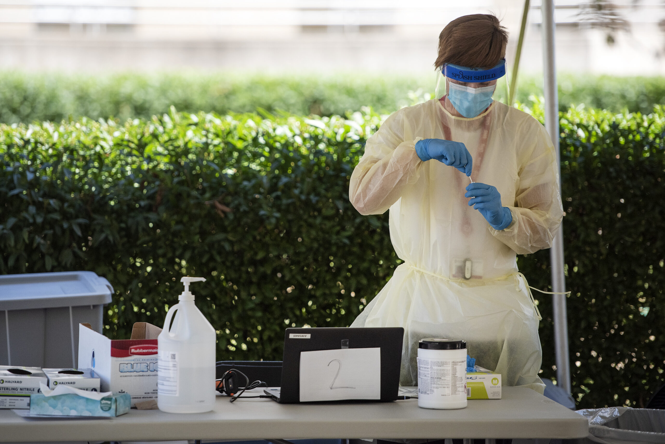 A man in a face mask, face shield, gown, and gloves handles a COVID-19 test at an outdoor testing facility under a tent